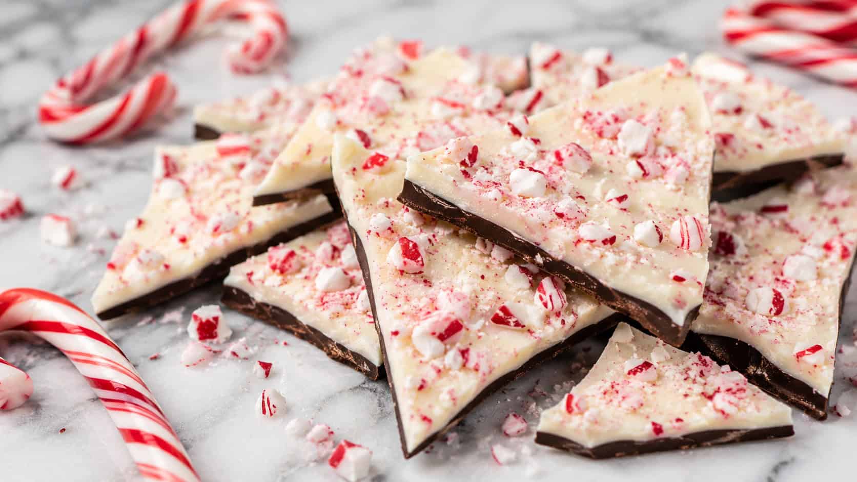 shards of peppermint bark with red and white candy cane bits on a white marble board in a wide horizontal shot, glossy two-tone chocolate, scattered candy canes, crisp natural light, shallow depth of field, landscape orientation