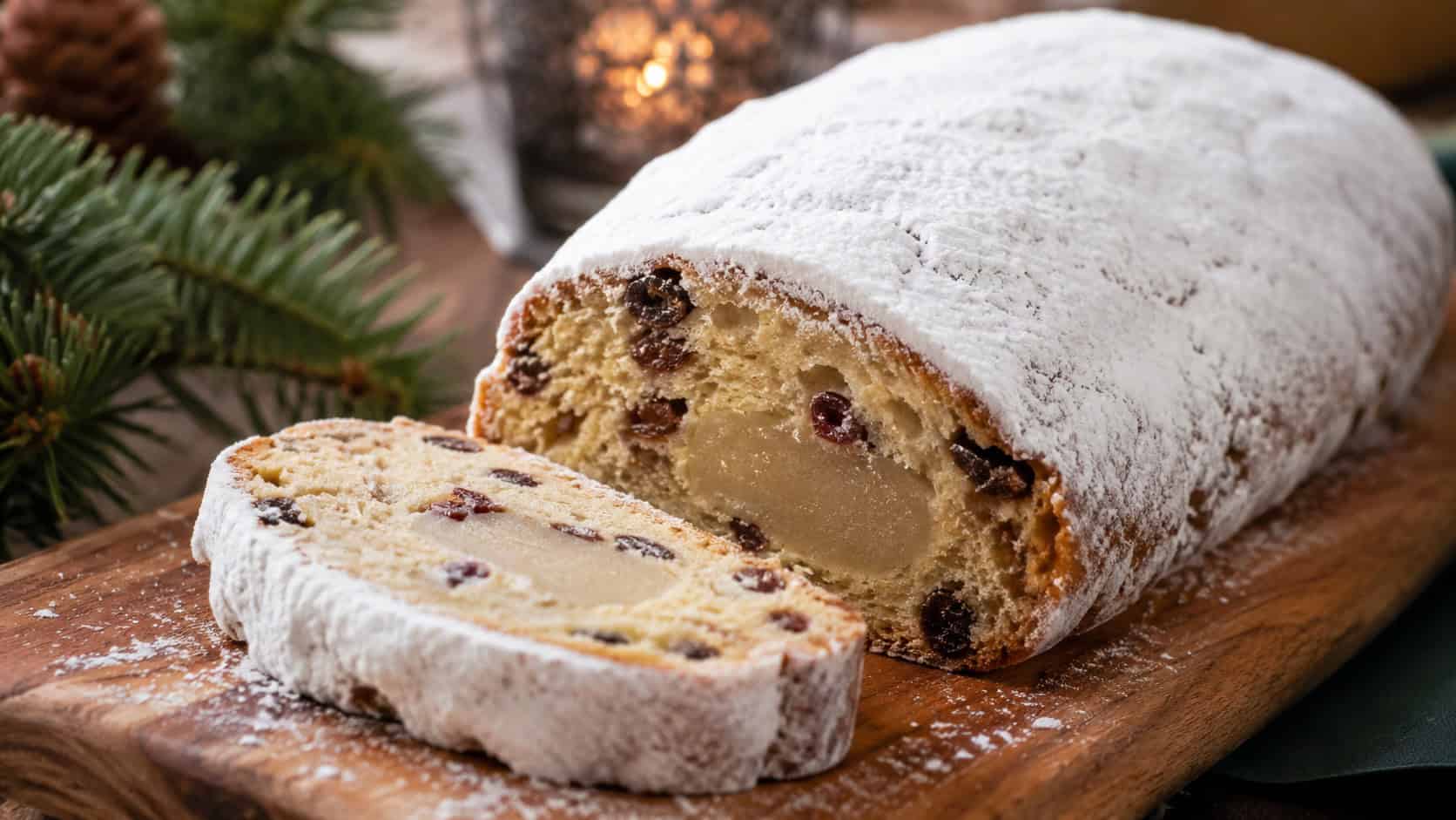a powdered-sugar-dusted stollen loaf with a slice cut to reveal fruit and marzipan on a wooden board in a wide horizontal shot, snowy sugar coating, holiday greenery nearby, warm natural light, shallow depth of field, landscape orientation