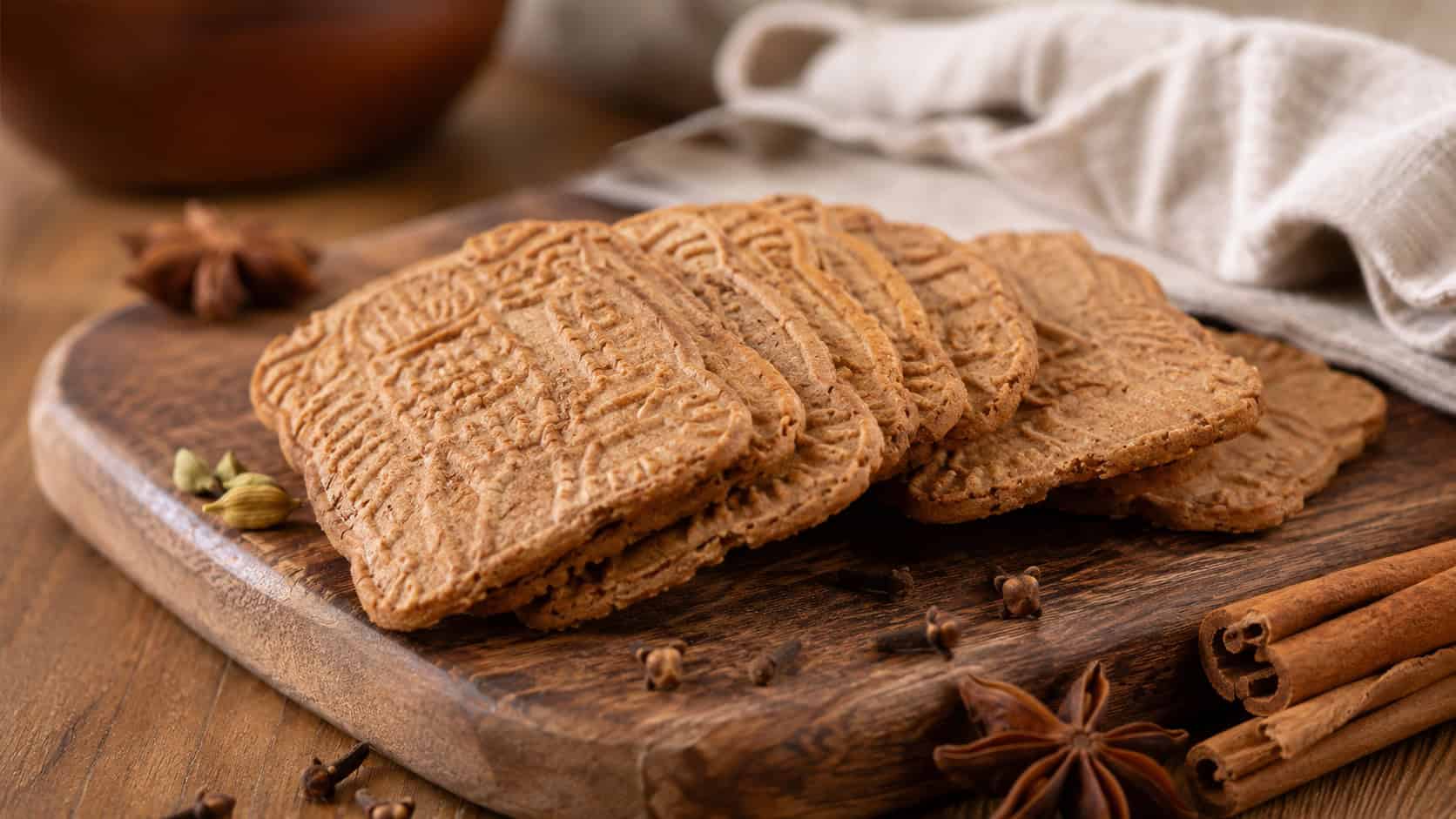 thin spiced speculaas cookies stacked and fanned on a wooden board in a wide horizontal shot, warm golden-brown color, scattered whole spices and a linen napkin, cozy natural light, shallow depth of field, landscape orientation
