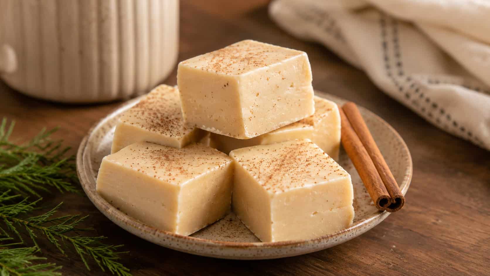pale eggnog fudge squares dusted with nutmeg stacked on a small plate in a wide horizontal shot, creamy smooth surface, a cinnamon stick and greenery nearby, warm natural light, shallow depth of field, landscape orientation