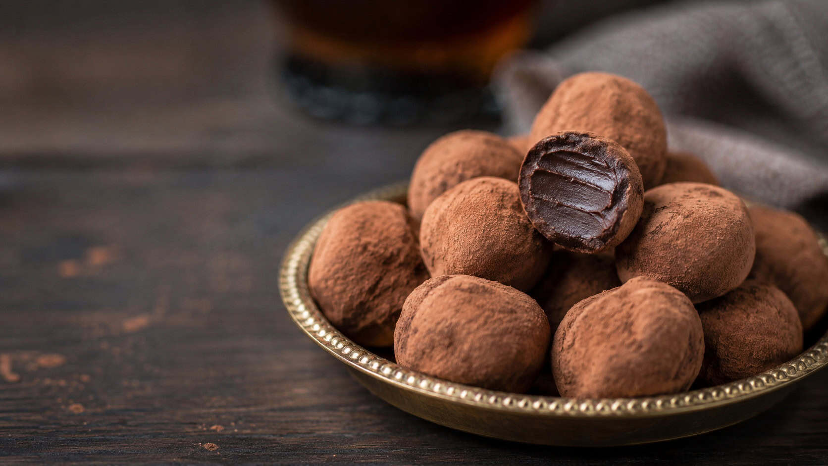 cocoa-dusted rum truffles piled in a small gold dish in a wide horizontal shot, one cut open to show the soft ganache center, dark moody table, soft natural light, shallow depth of field, landscape orientation