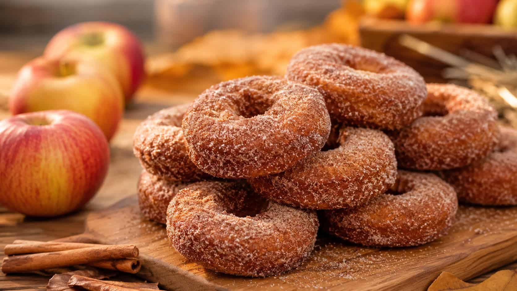 cinnamon-sugar apple cider donuts stacked on a wooden board in a wide horizontal shot, crackly sugary coating, fresh apples and cinnamon sticks nearby, warm autumn natural light, shallow depth of field, landscape orientation