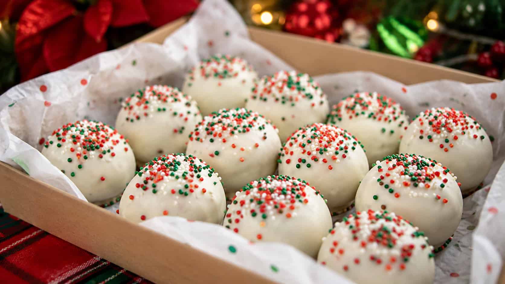 round white-chocolate-coated cookie truffles topped with red and green sprinkles, in a wide horizontal shot in a paper-lined gift box on a holiday table, bright festive light, shallow depth of field, landscape orientation