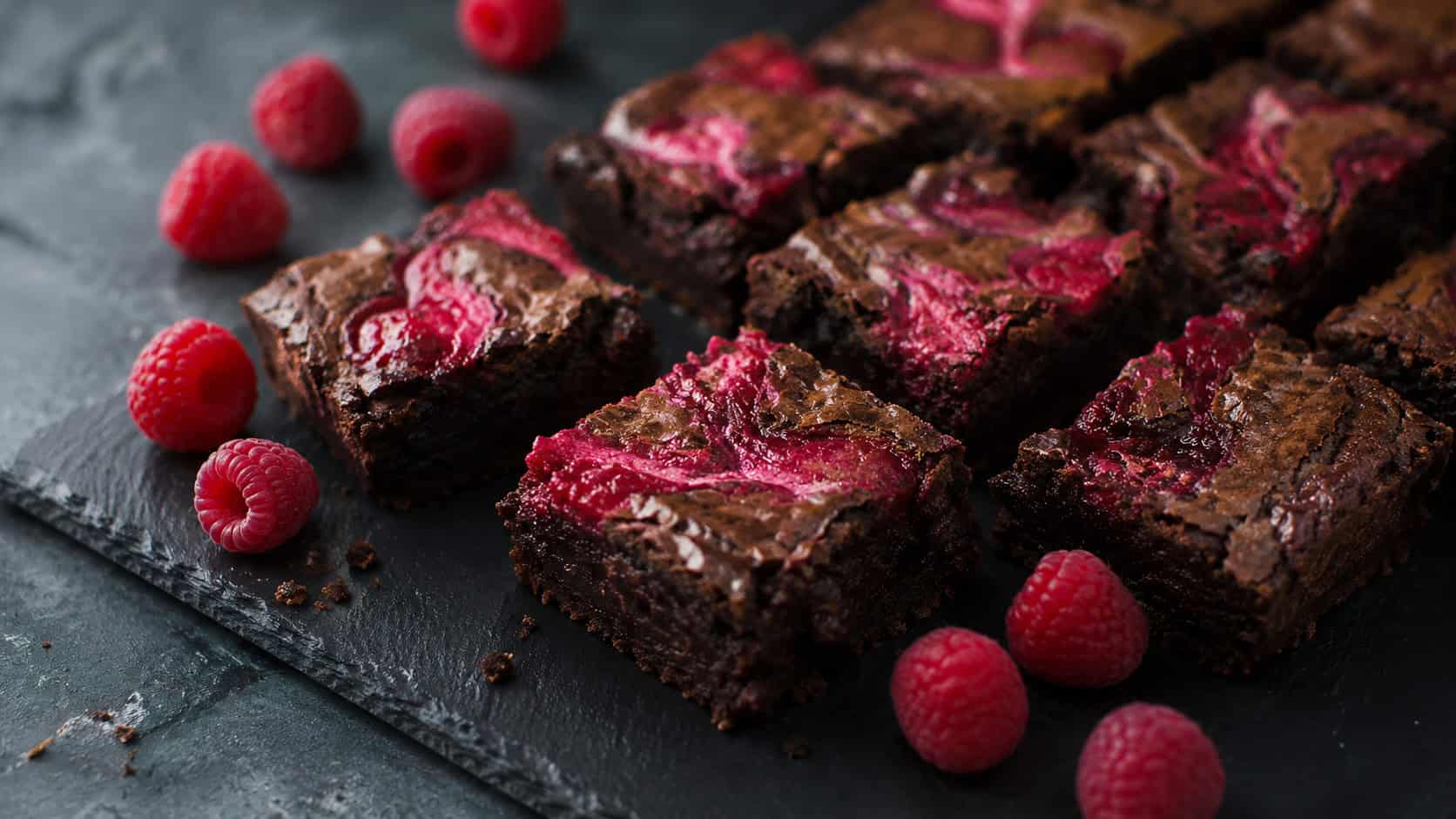 fudgy brownies with bright raspberry swirls on top cut into squares, in a wide horizontal shot on a dark slate board with fresh raspberries scattered around, moody natural light, shallow depth of field, landscape orientation