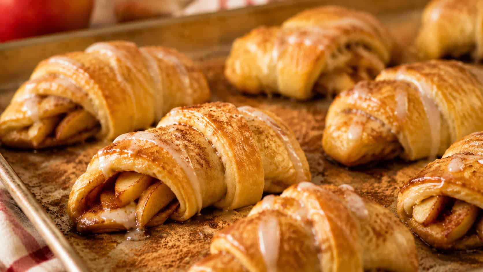 golden baked crescent rolls filled with cinnamon apple slices drizzled with icing, in a wide horizontal shot on a baking sheet with a dusting of cinnamon, warm cozy light, shallow depth of field, landscape orientation