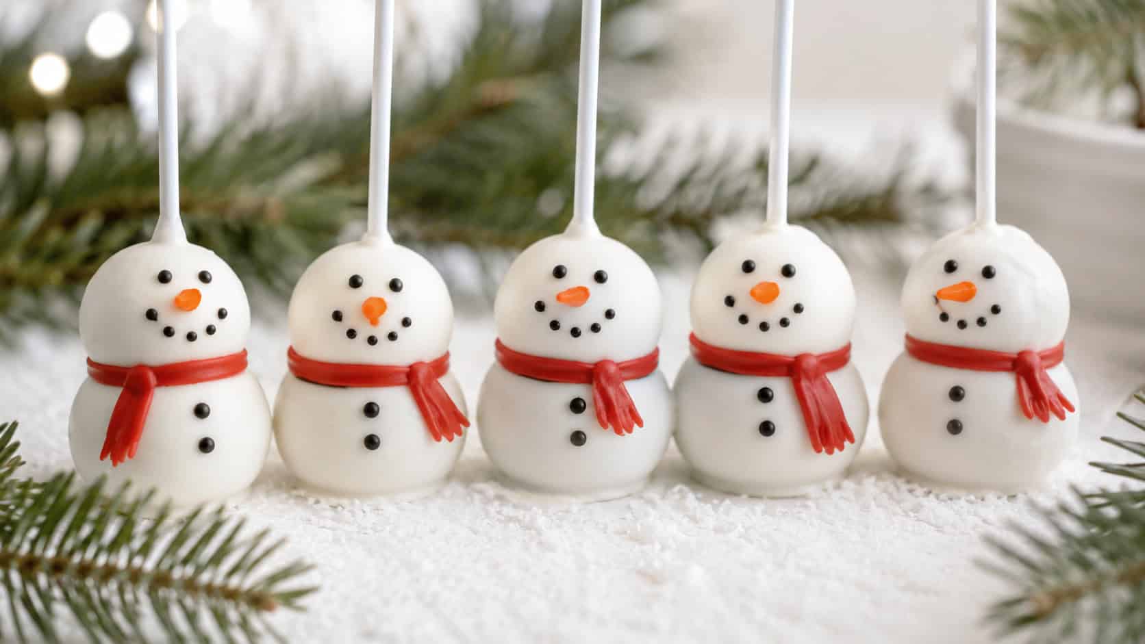 white cake pops decorated as cheerful little snowmen with tiny faces, in a wide horizontal shot standing in a row on a snowy white surface with greenery, bright cheerful light, shallow depth of field, landscape orientation
