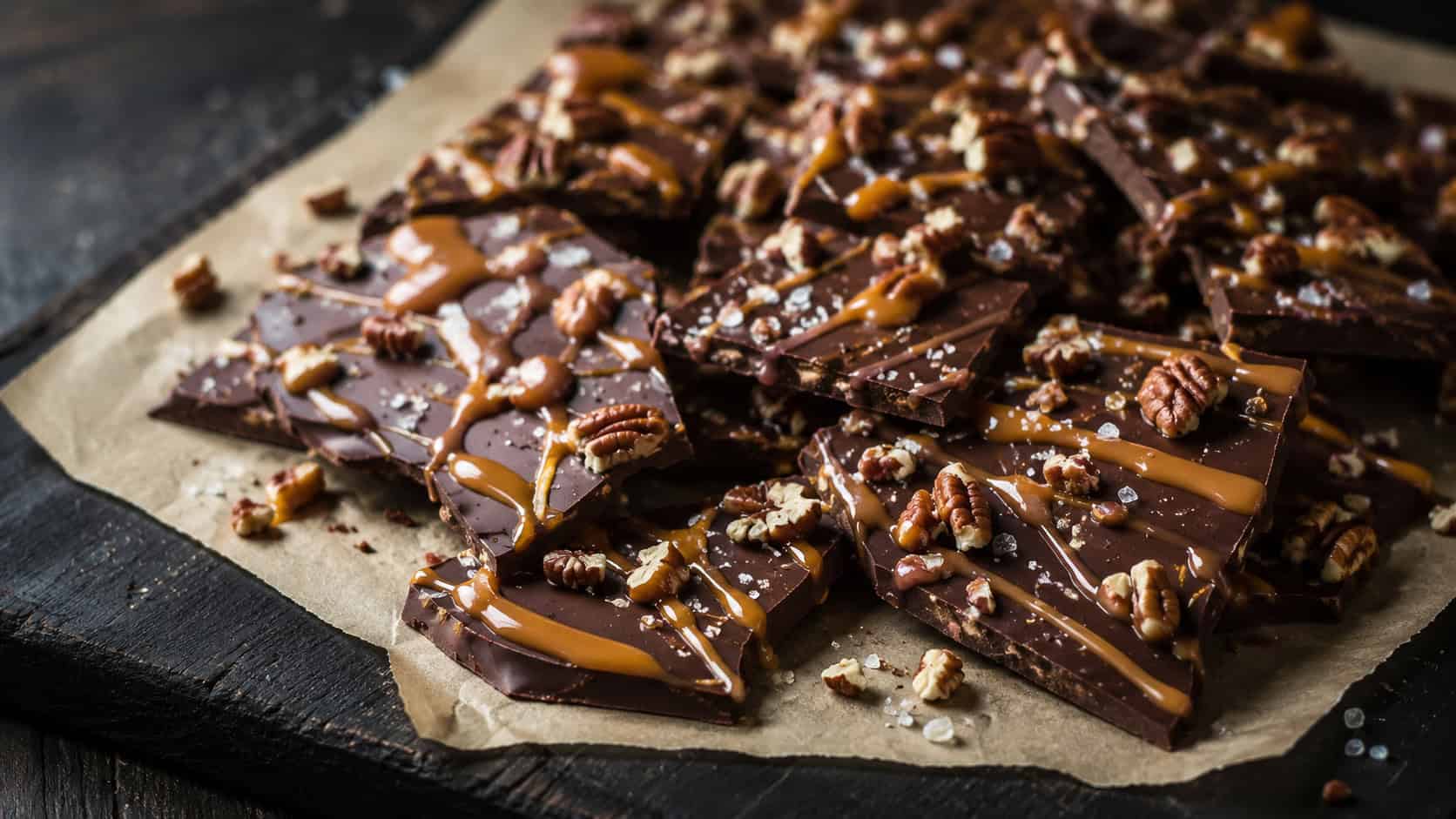broken pieces of dark chocolate bark drizzled with caramel and flecked with sea salt and pecans, in a wide horizontal shot on parchment over a dark board, dramatic natural light, shallow depth of field, landscape orientation