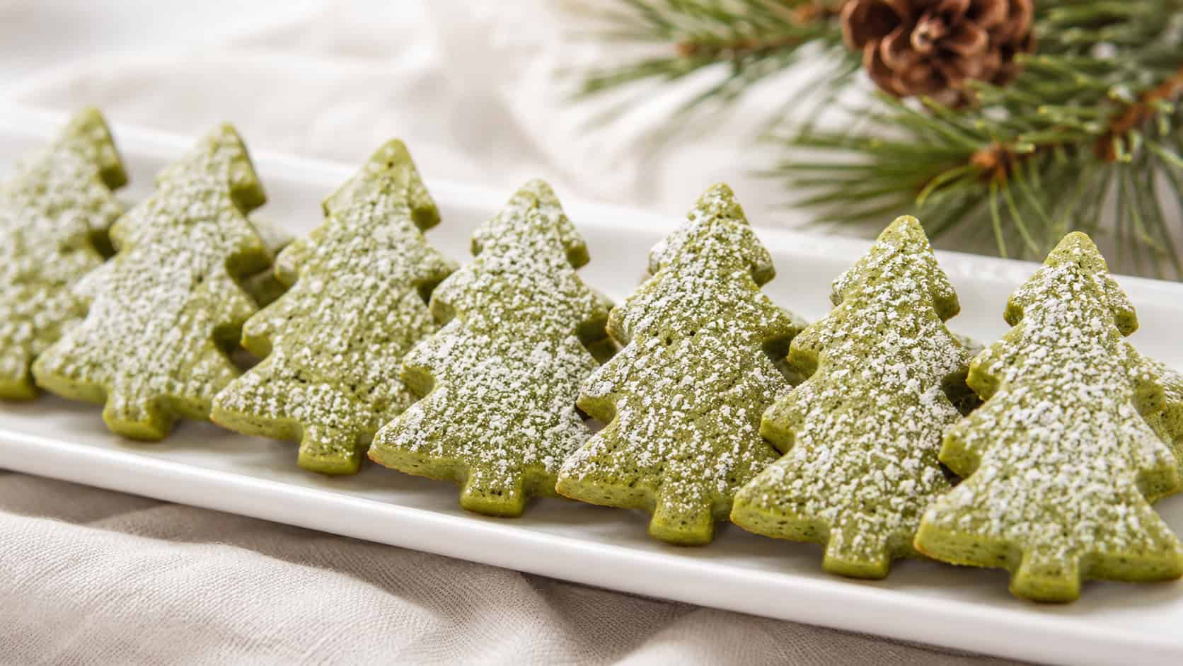 green matcha shortbread cookies shaped like Christmas trees dusted with powdered sugar, in a wide horizontal shot lined up on a white plate with a sprig of pine, bright clean light, shallow depth of field, landscape orientation