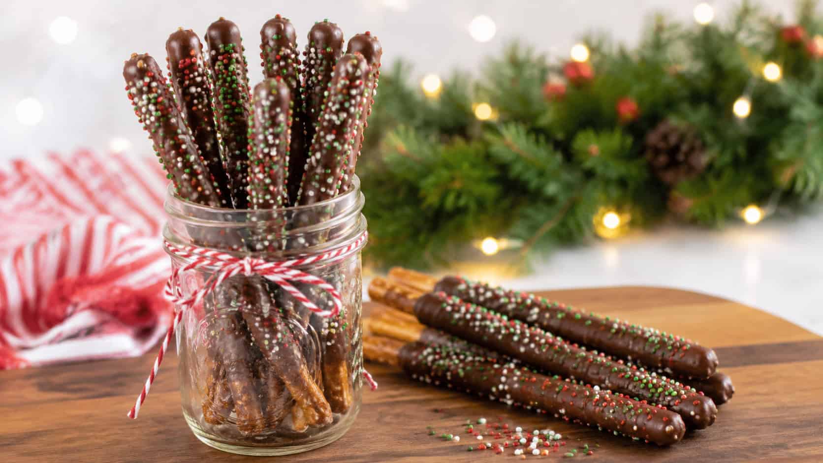 chocolate-dipped pretzel rods covered in red and green sprinkles, in a wide horizontal shot arranged in a mason jar and laid across a wooden board, bright cheerful light, shallow depth of field, landscape orientation