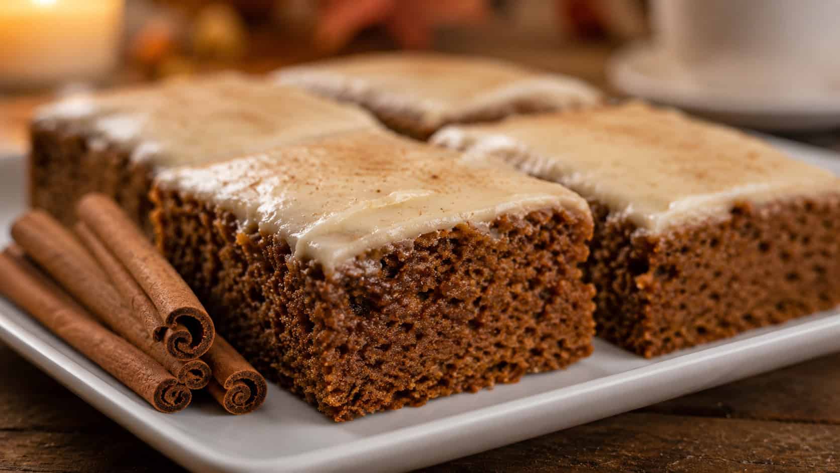 frosted gingerbread bars cut into squares showing the dark spiced interior, in a wide horizontal shot on a white plate with cinnamon sticks alongside, warm cozy light, shallow depth of field, landscape orientation
