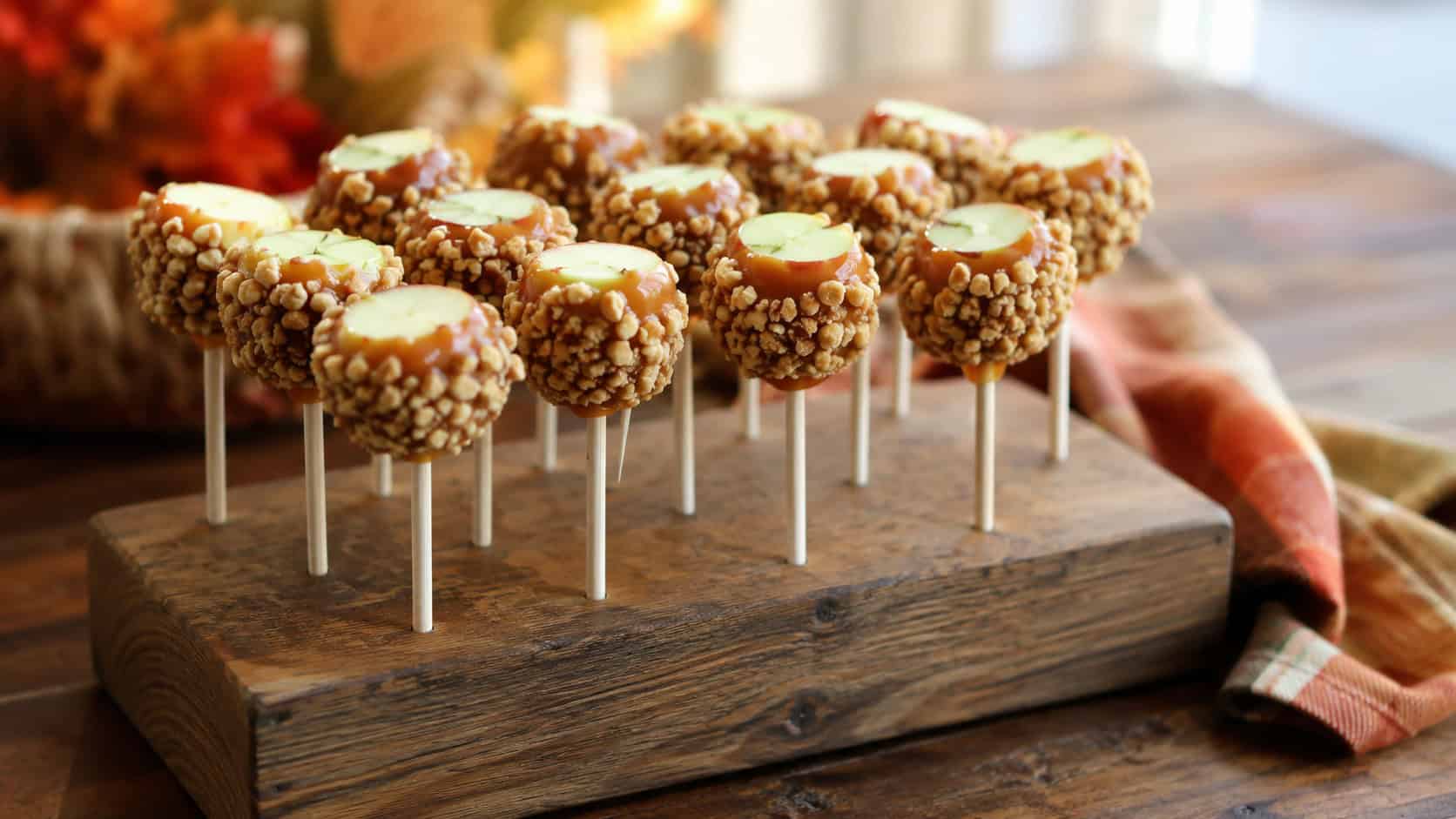 mini caramel-dipped apple bites on sticks rolled in crushed nuts, in a wide horizontal shot standing in a block or jar on a rustic table with autumn-toned napkins, warm natural light, shallow depth of field, landscape orientation