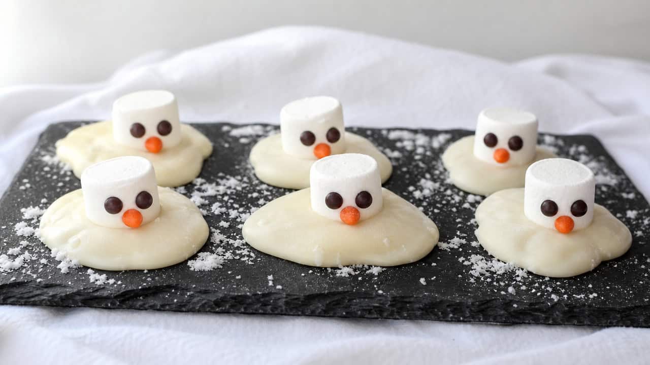 adorable melting snowman cookie truffles coated in white chocolate with marshmallow heads, chocolate chip eyes, and orange candy noses, arranged in a wide horizontal shot on a slate board over a snowy white cloth, soft natural light, shallow depth of field, landscape orientation