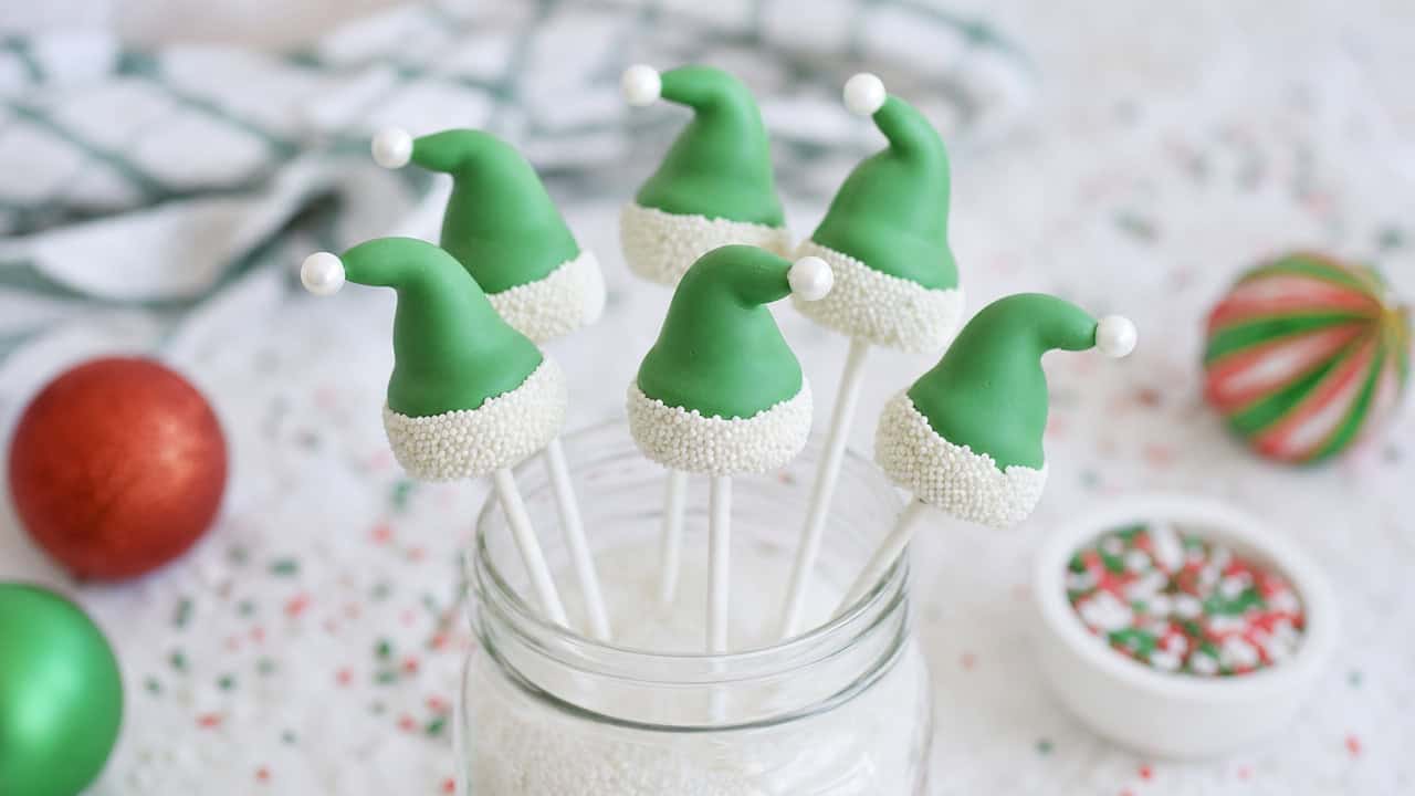 playful green elf hat marshmallow pops with white sprinkle brims and candy pearl tips standing upright in a jar, shown in a wide horizontal composition on a snowy white surface with scattered sprinkles, bright natural light, shallow depth of field, landscape orientation