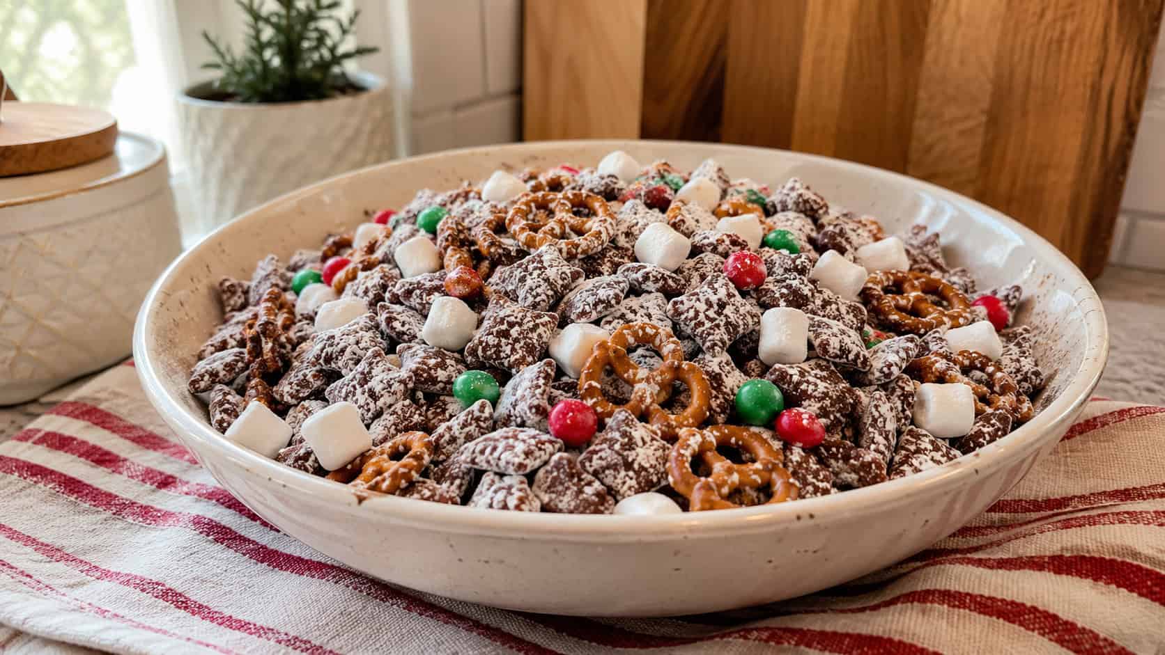 a generous bowl of chocolate-coated cereal snack mix dusted in powdered sugar with pretzels, mini marshmallows, and red and green candies, shown in a wide horizontal shot on a cozy kitchen counter with a striped napkin, warm natural light, shallow depth of field, landscape orientation