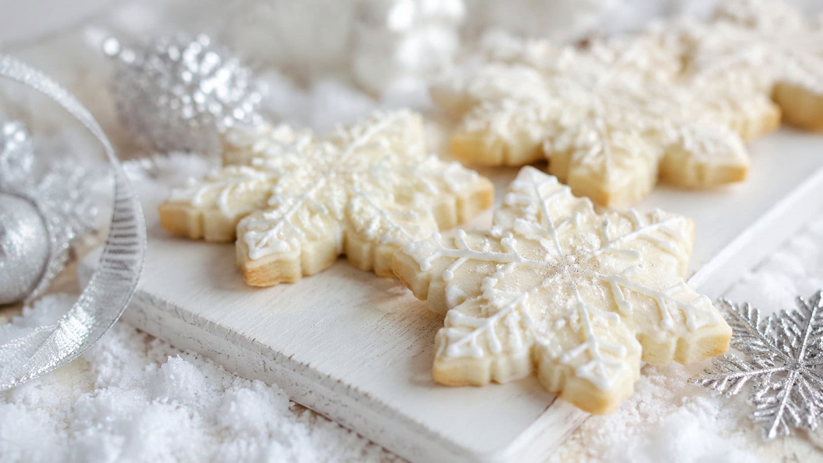 white-iced snowflake sugar cookies dusted with sparkling sugar on a pale board in a wide horizontal shot, faux snow and silver ribbon nearby, soft bright light, shallow depth of field, landscape orientation