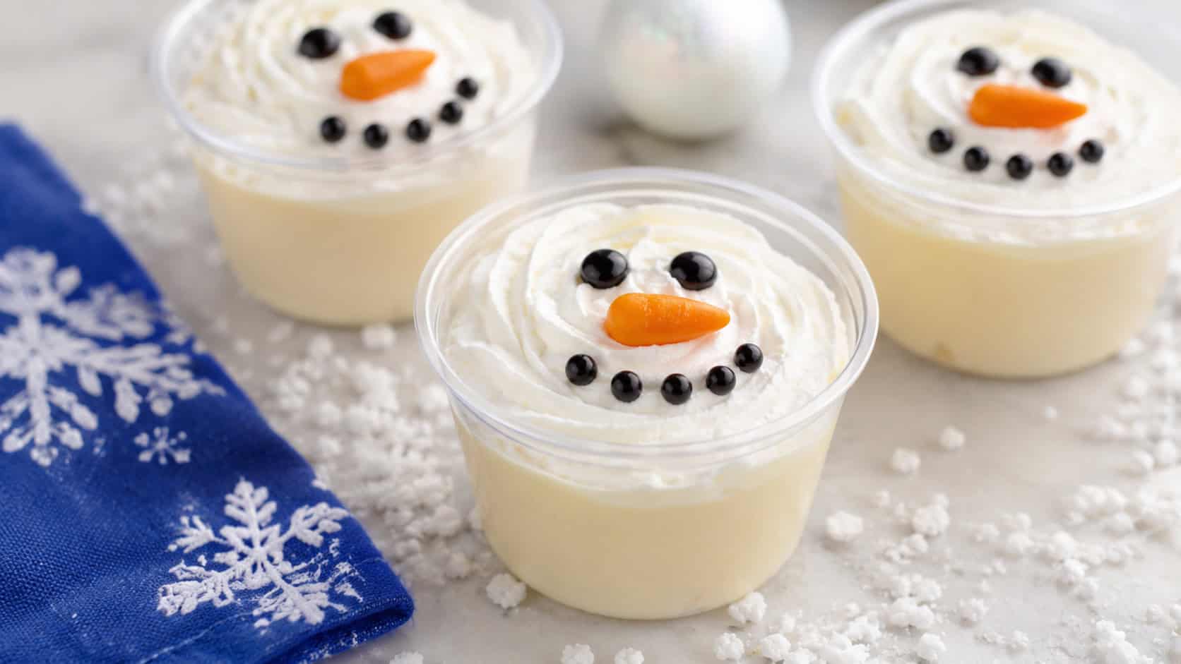 clear pudding cups topped with whipped cream snowman faces with candy features on a white table in a wide horizontal shot, faux snow and a blue napkin nearby, soft bright light, shallow depth of field, landscape orientation