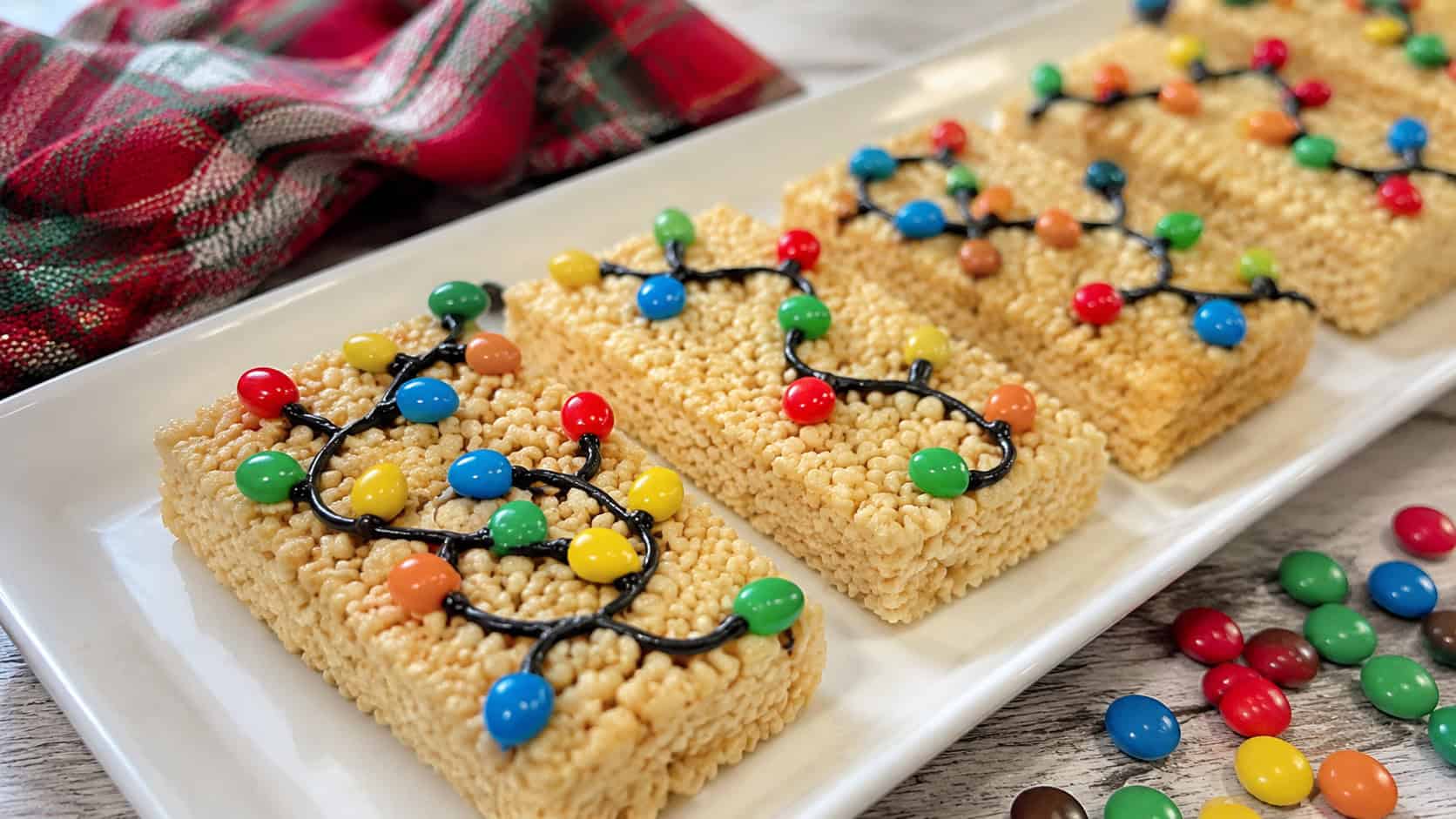 crispy cereal treat bars decorated with a black icing wire and colorful candy lights on a white plate in a wide horizontal shot, a festive cloth and loose candies nearby, bright natural light, shallow depth of field, landscape orientation
