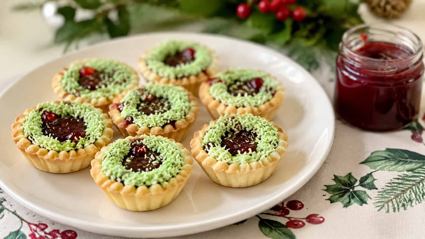 mini jam tarts topped with green-tinted coconut tinsel on a white plate in a wide horizontal shot, a holiday cloth and a small jar of jam nearby, soft bright light, shallow depth of field, landscape orientation