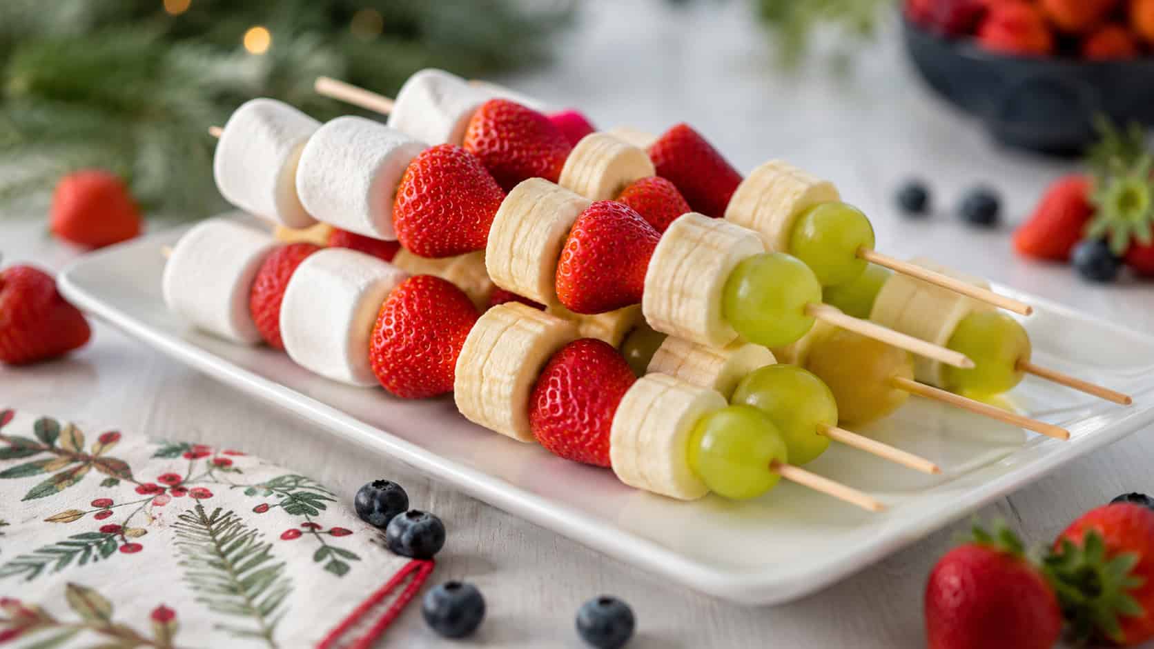 fruit skewers stacked with green grapes, banana, strawberry, and a marshmallow on top arranged on a white platter in a wide horizontal shot, scattered berries and a holiday napkin nearby, bright natural light, shallow depth of field, landscape orientation