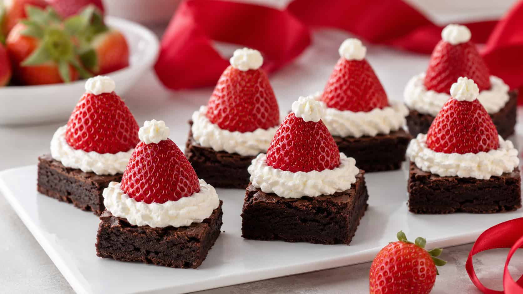 fudgy brownie squares each topped with a strawberry and whipped cream to look like Santa hats on a white plate in a wide horizontal shot, fresh strawberries and a red ribbon nearby, soft bright light, shallow depth of field, landscape orientation