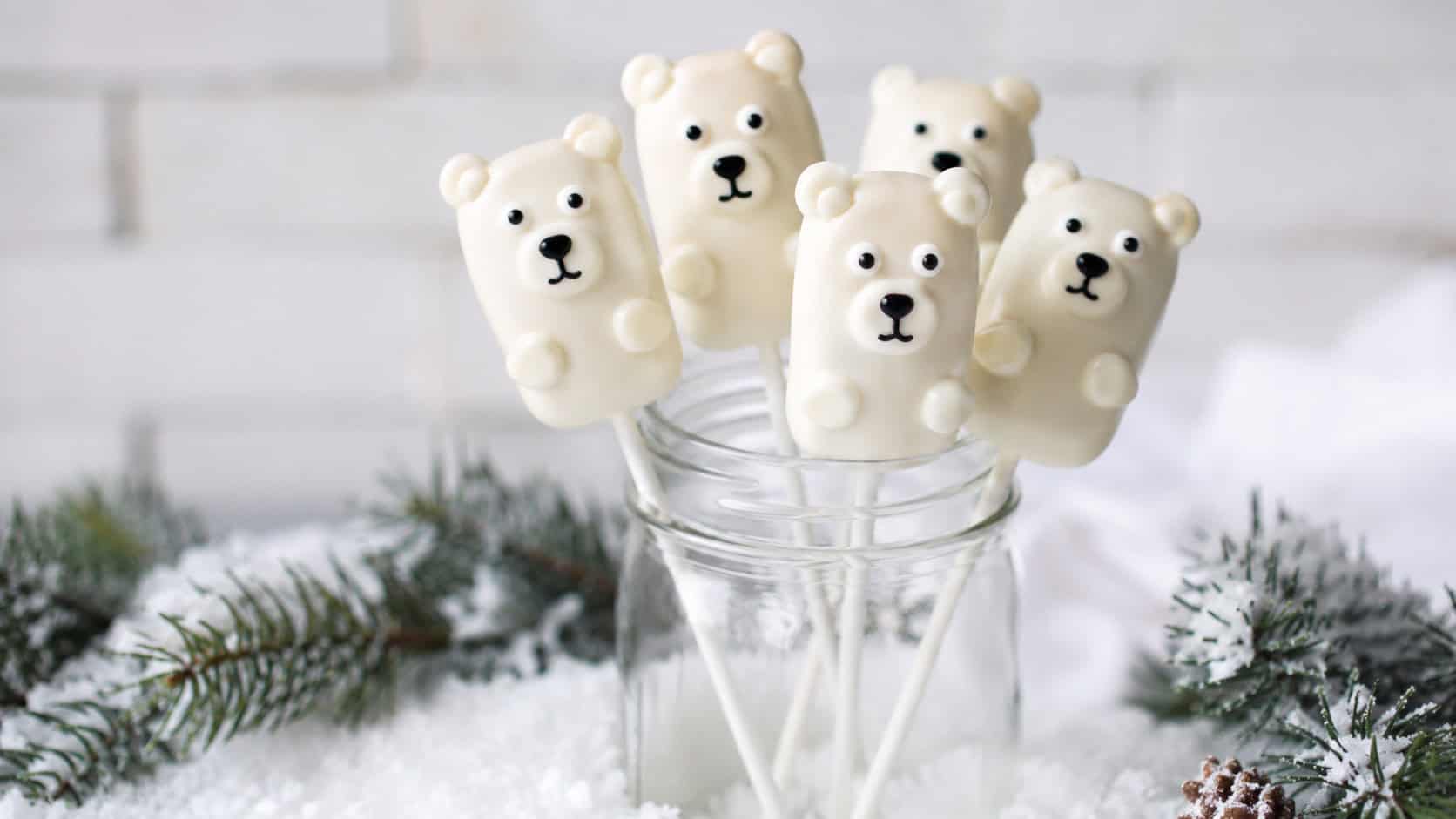 white-chocolate-dipped marshmallow polar bear pops with candy eyes and ears standing upright in a jar in a wide horizontal shot, faux snow and pine sprigs around the base, soft bright light, shallow depth of field, landscape orientation