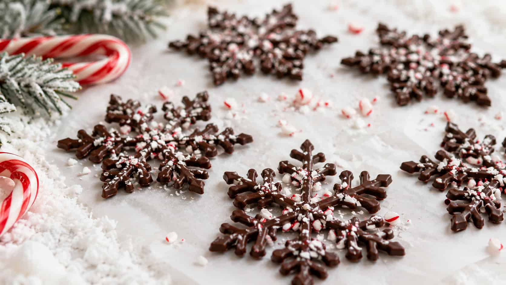 delicate chocolate snowflake shapes dusted with crushed peppermint arranged on white parchment in a wide horizontal shot, candy canes and faux snow nearby, soft bright light, shallow depth of field, landscape orientation