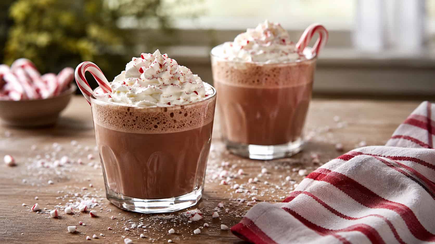 two glasses of chocolate milk topped with whipped cream and candy cane stirrers on a wooden table in a wide horizontal shot, scattered crushed peppermint and a striped napkin nearby, soft natural light, shallow depth of field, landscape orientation