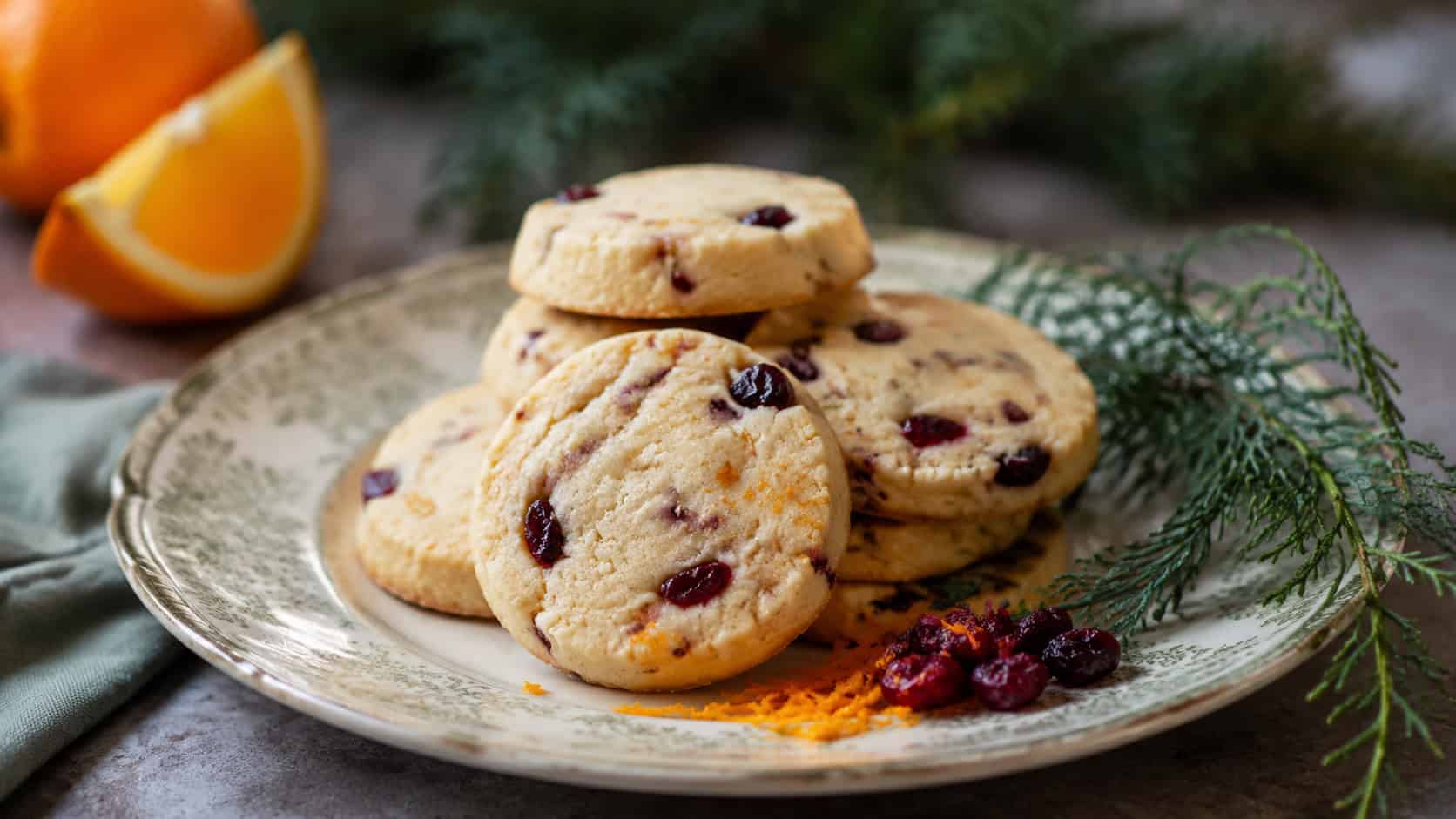 gluten-free cranberry orange shortbread rounds in a wide horizontal shot, stacked on a vintage plate with orange zest and dried cranberries beside a sprig of greenery, natural light, shallow depth of field, landscape orientation