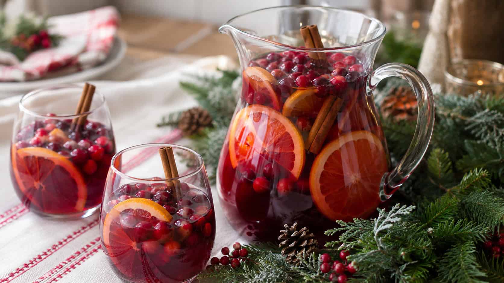 a glass pitcher of red holiday sangria with floating cranberries and orange slices in a wide horizontal shot, cinnamon sticks and filled glasses, on a festive table with greenery, bright natural light, shallow depth of field, landscape orientation