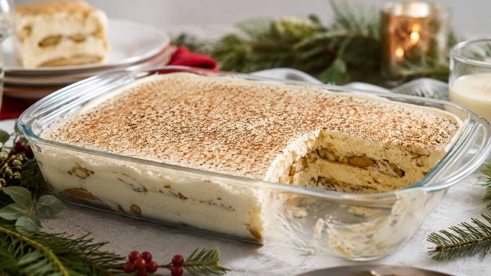 eggnog tiramisu in a glass dish in a wide horizontal shot, creamy layers and a dusting of nutmeg, one portion scooped out, on a festive table with greenery, soft natural light, shallow depth of field, landscape orientation