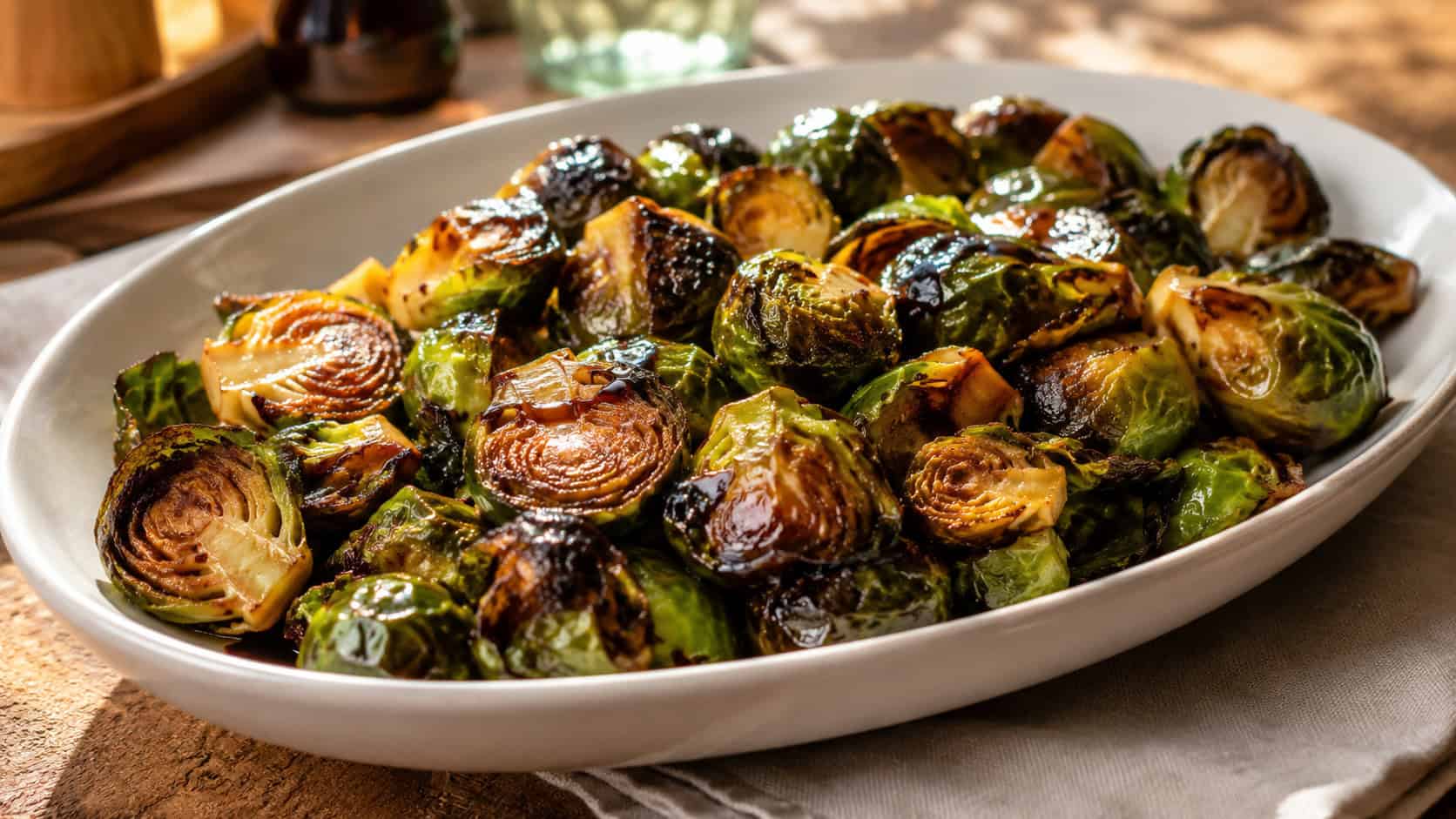 crispy caramelized brussels sprouts glazed in balsamic glistening in a wide horizontal shot in a white serving dish on a rustic table with a linen napkin, warm natural light, shallow depth of field, landscape orientation