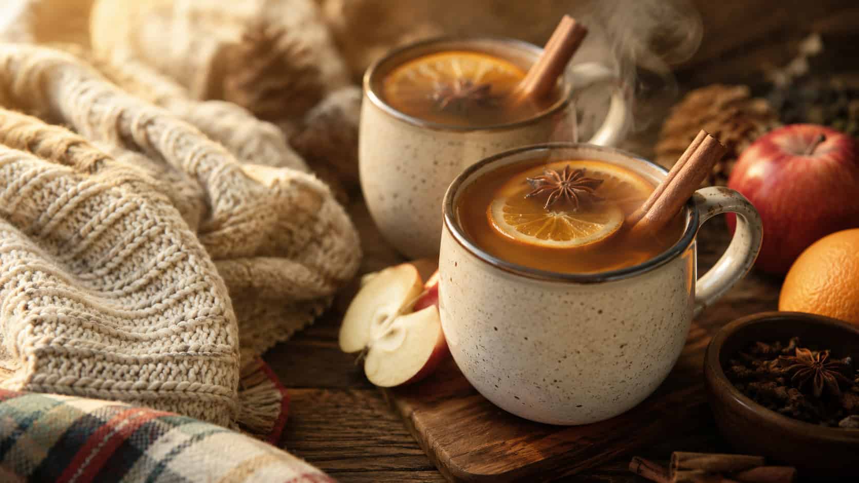 steaming mulled apple cider in mugs with cinnamon sticks, orange slices, and star anise floating on top, shown in a wide horizontal composition on a cozy wooden table beside a knit blanket, warm golden light, shallow depth of field, landscape orientation