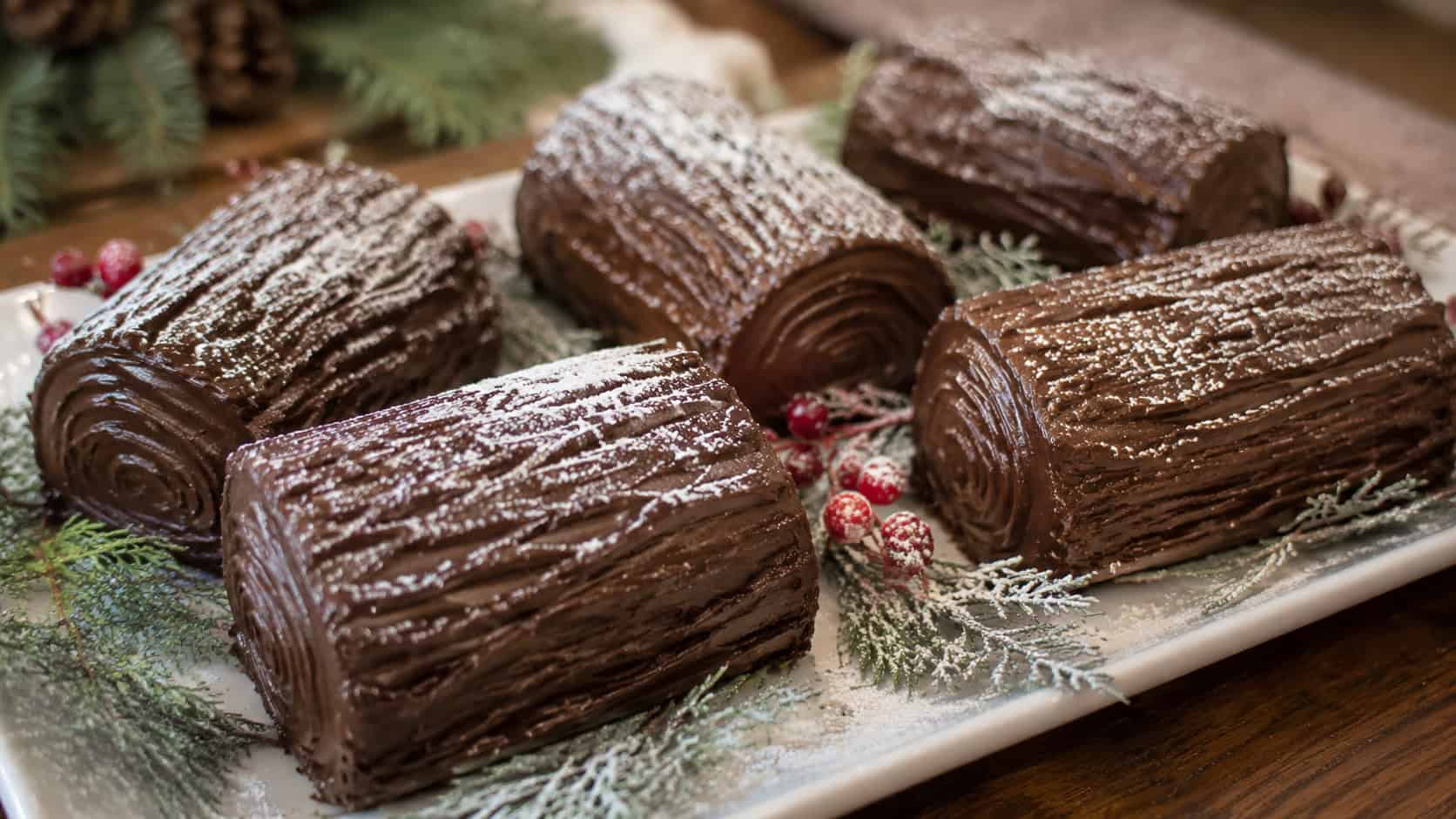 mini chocolate yule log cakes frosted with bark-textured chocolate frosting and dusted with powdered sugar snow, arranged in a wide horizontal shot on a white plate with greenery and berries, warm natural light, shallow depth of field, landscape orientation