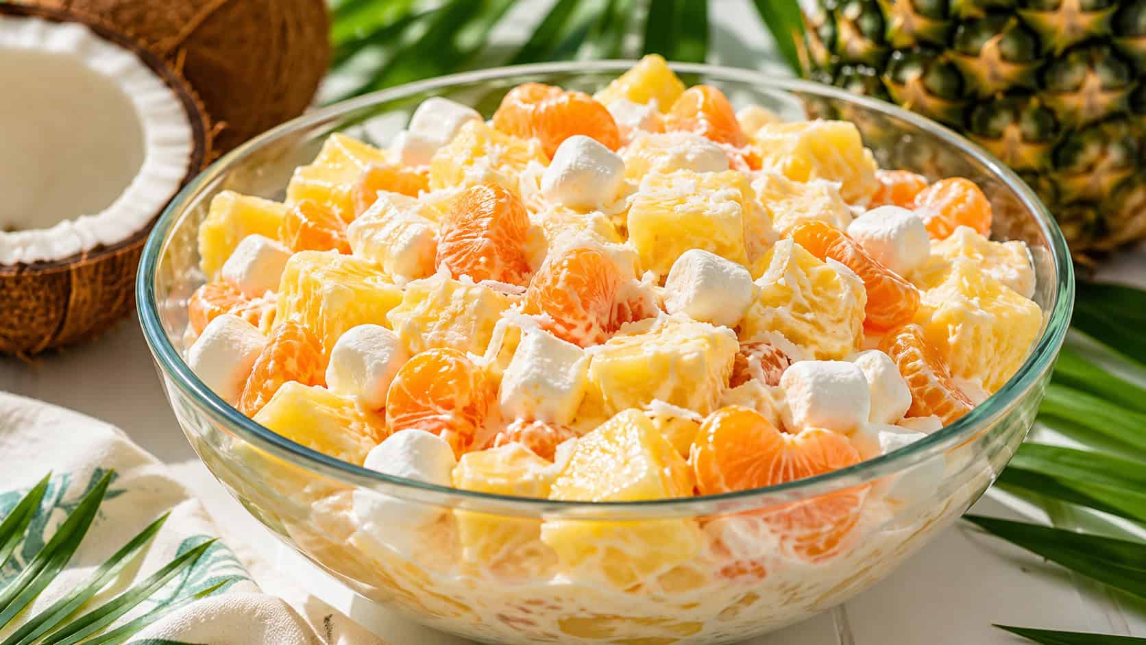 a bright pineapple and coconut salad with mandarin oranges and marshmallows in a creamy dressing, served in a wide horizontal shot in a glass bowl on a white table with tropical greenery nearby, fresh natural light, shallow depth of field, landscape orientation