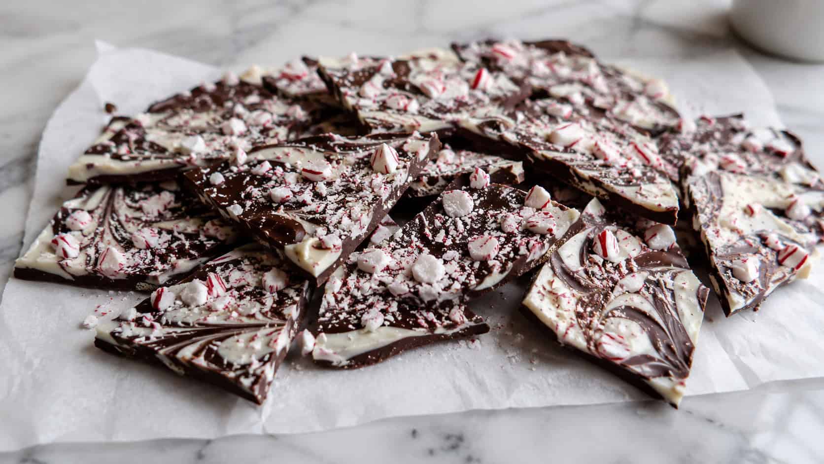 swirled dark and white chocolate bark topped with crushed red and white peppermint candies broken into shards, arranged in a wide horizontal shot on parchment over a marble counter, cool natural light, shallow depth of field, landscape orientation