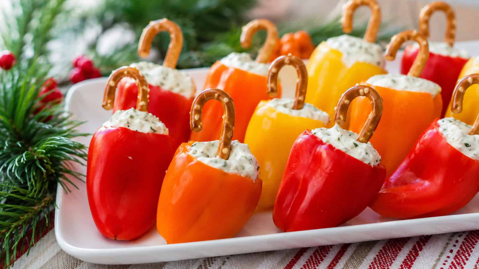 colorful stuffed mini peppers arranged on a white platter in a wide horizontal shot, red, orange, and yellow peppers filled with creamy cheese and topped with pretzel hooks, festive greenery alongside, bright fresh light, shallow depth of field, landscape orientation