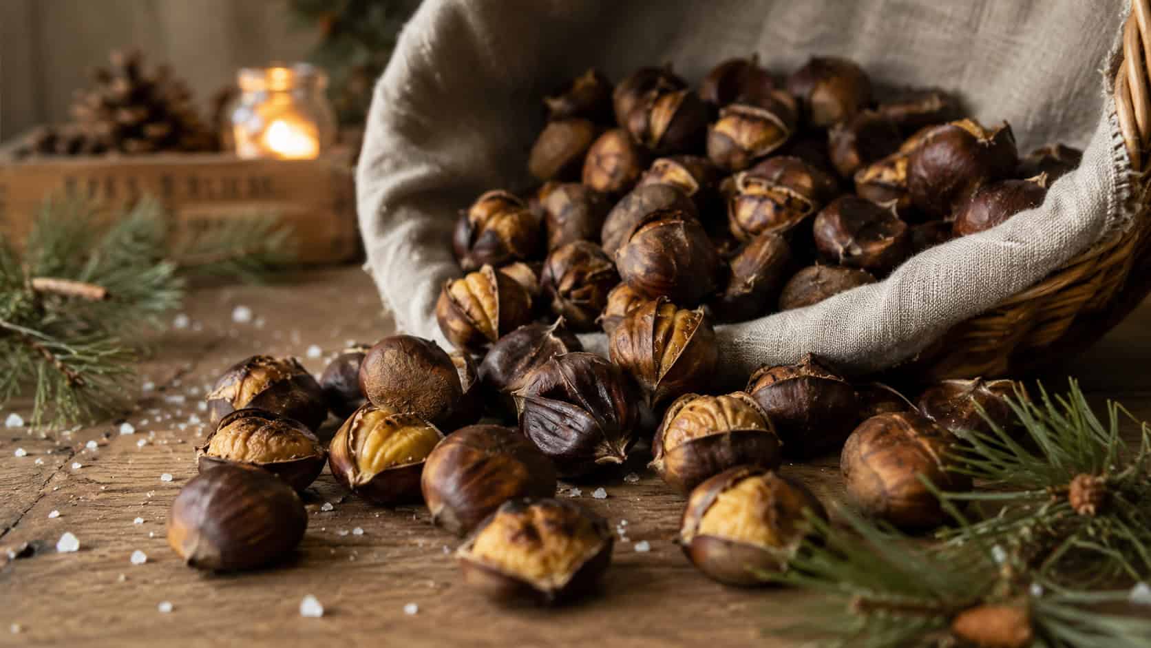 roasted chestnuts tumbling from a linen-lined basket onto a wooden table in a wide horizontal shot, split brown shells revealing creamy centers, sea salt and pine sprigs scattered around, cozy golden light, shallow depth of field, landscape orientation