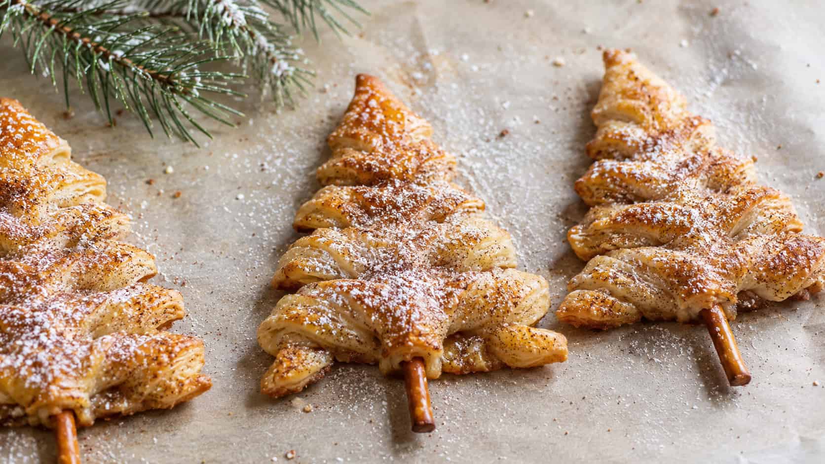 cinnamon sugar puff pastry trees lined up on parchment in a wide horizontal shot, flaky golden triangles glistening with cinnamon sugar and pretzel trunks, a dusting of powdered sugar and pine sprigs nearby, bright natural light, shallow depth of field, landscape orientation