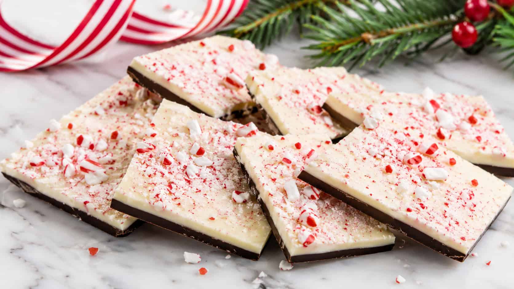 peppermint bark pieces fanned across a marble slab in a wide horizontal shot, layered dark and white chocolate sprinkled with red and white candy bits, a striped ribbon and pine beside them, bright clean light, shallow depth of field, landscape orientation