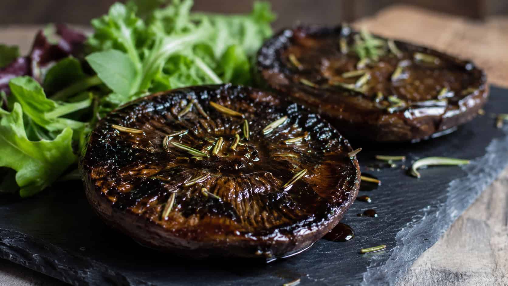 roasted portobello mushroom steaks brushed with glossy dark balsamic glaze and sprinkled with rosemary, in a wide horizontal shot on a slate plate with greens alongside, moody natural light, shallow depth of field, landscape orientation