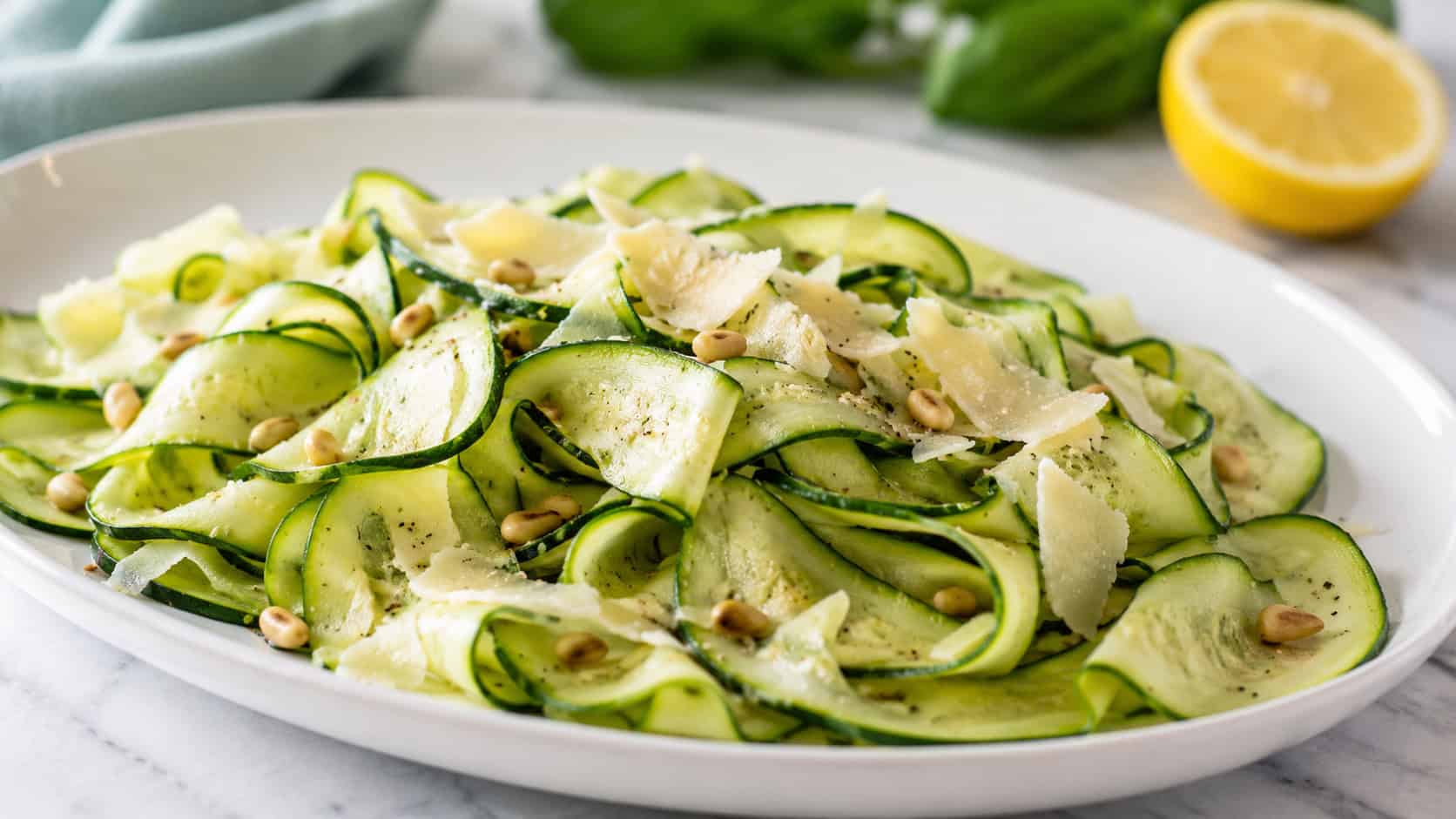 delicate raw zucchini ribbons tossed with shaved parmesan and pine nuts, in a wide horizontal shot piled on a white platter with a lemon half nearby, bright fresh light, shallow depth of field, landscape orientation