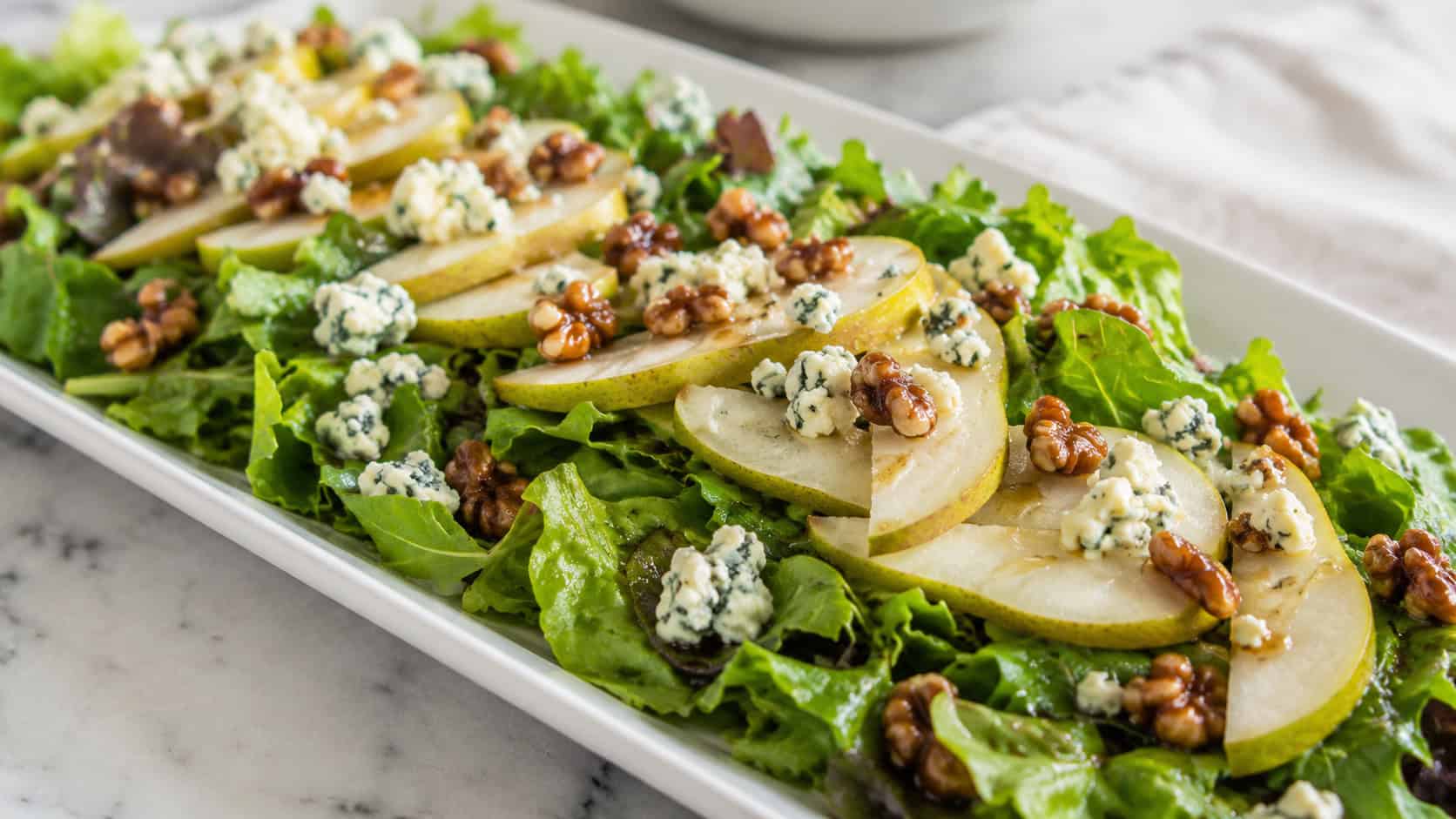 a fresh green salad topped with sliced pears, crumbled blue cheese, and candied walnuts, in a wide horizontal shot on a long white platter with a drizzle of dressing, bright natural light, shallow depth of field, landscape orientation