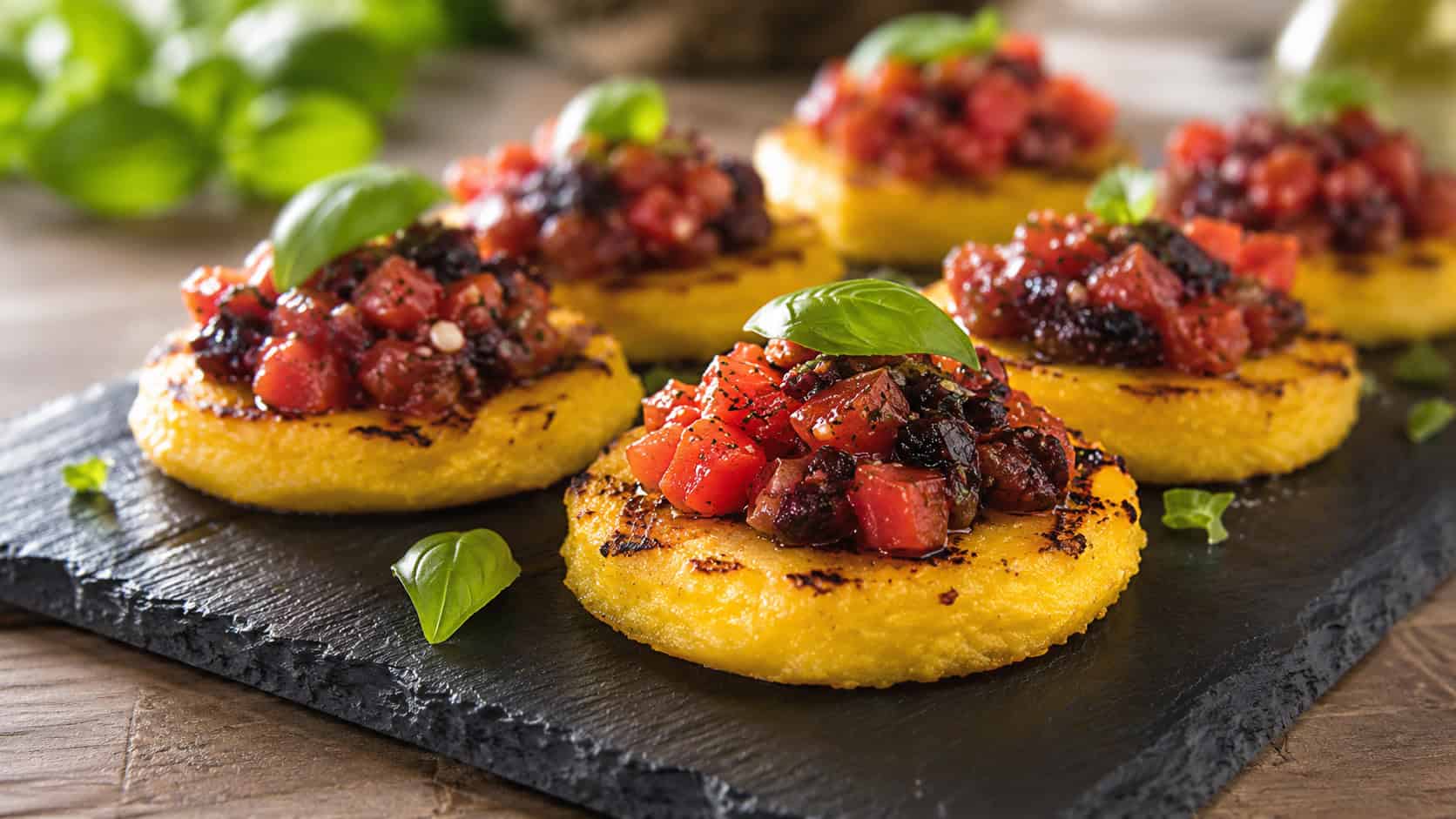 crisp golden grilled polenta rounds topped with red tomato and dark olive tapenade, in a wide horizontal shot on a slate board garnished with basil, warm natural light, shallow depth of field, landscape orientation