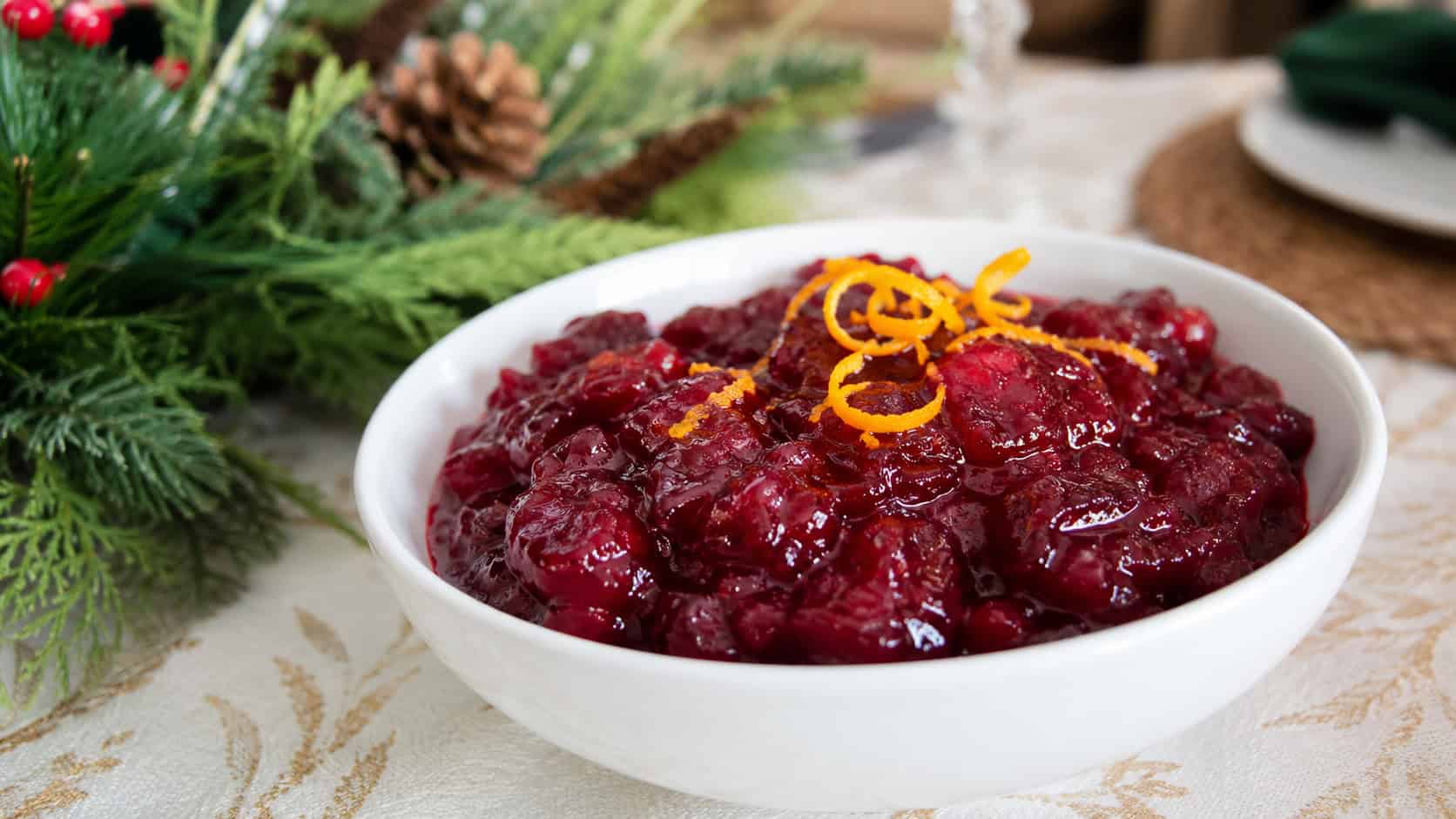 glossy ruby orange-cranberry sauce in a small white bowl with curls of orange zest in a wide horizontal shot, on a holiday table beside greenery, natural light, shallow depth of field, landscape orientation