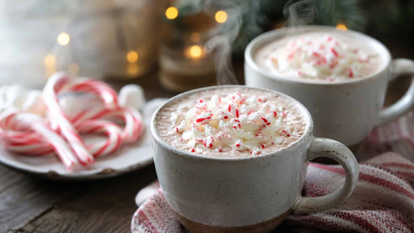 creamy peppermint hot chocolate in mugs topped with whipped cream and crushed candy canes in a wide horizontal shot, steam rising, cozy table with candy canes and lights blurred behind, natural light, shallow depth of field, landscape orientation