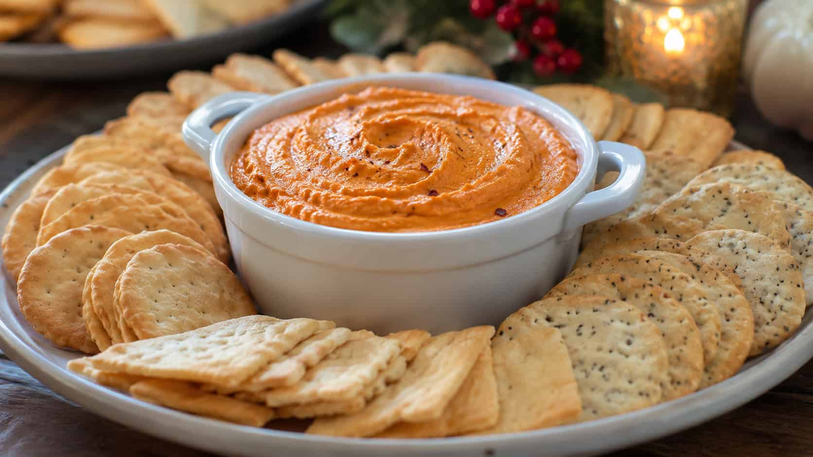 warm creamy roasted red pepper dip in a small crock with a swirl on top and crackers fanned around it in a wide horizontal shot, on a festive appetizer table, natural light, shallow depth of field, landscape orientation