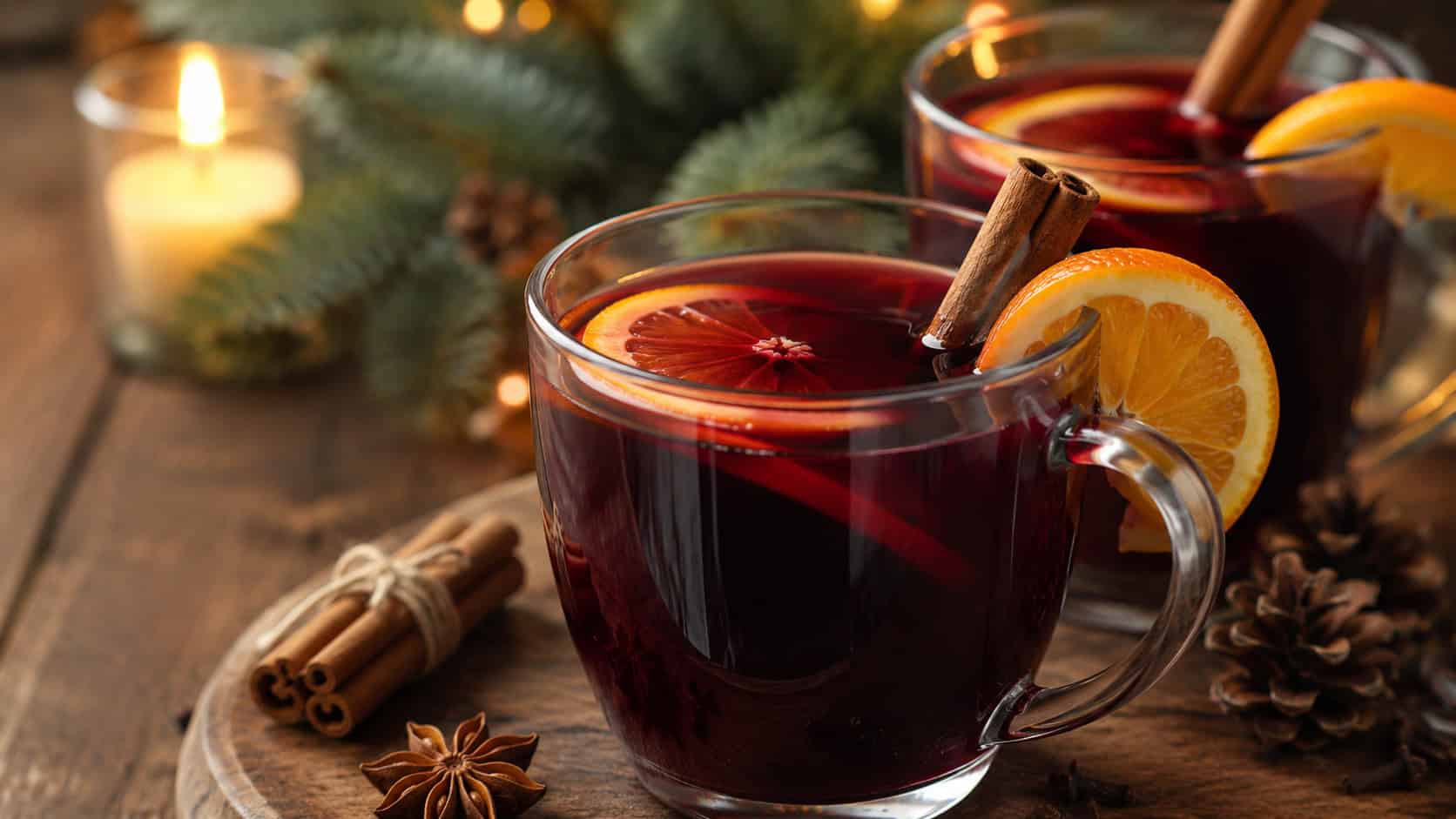 warm mulled wine in glass mugs garnished with orange slices and cinnamon sticks in a wide horizontal shot, deep ruby color, cozy table with candles and pine softly blurred behind, natural light, shallow depth of field, landscape orientation