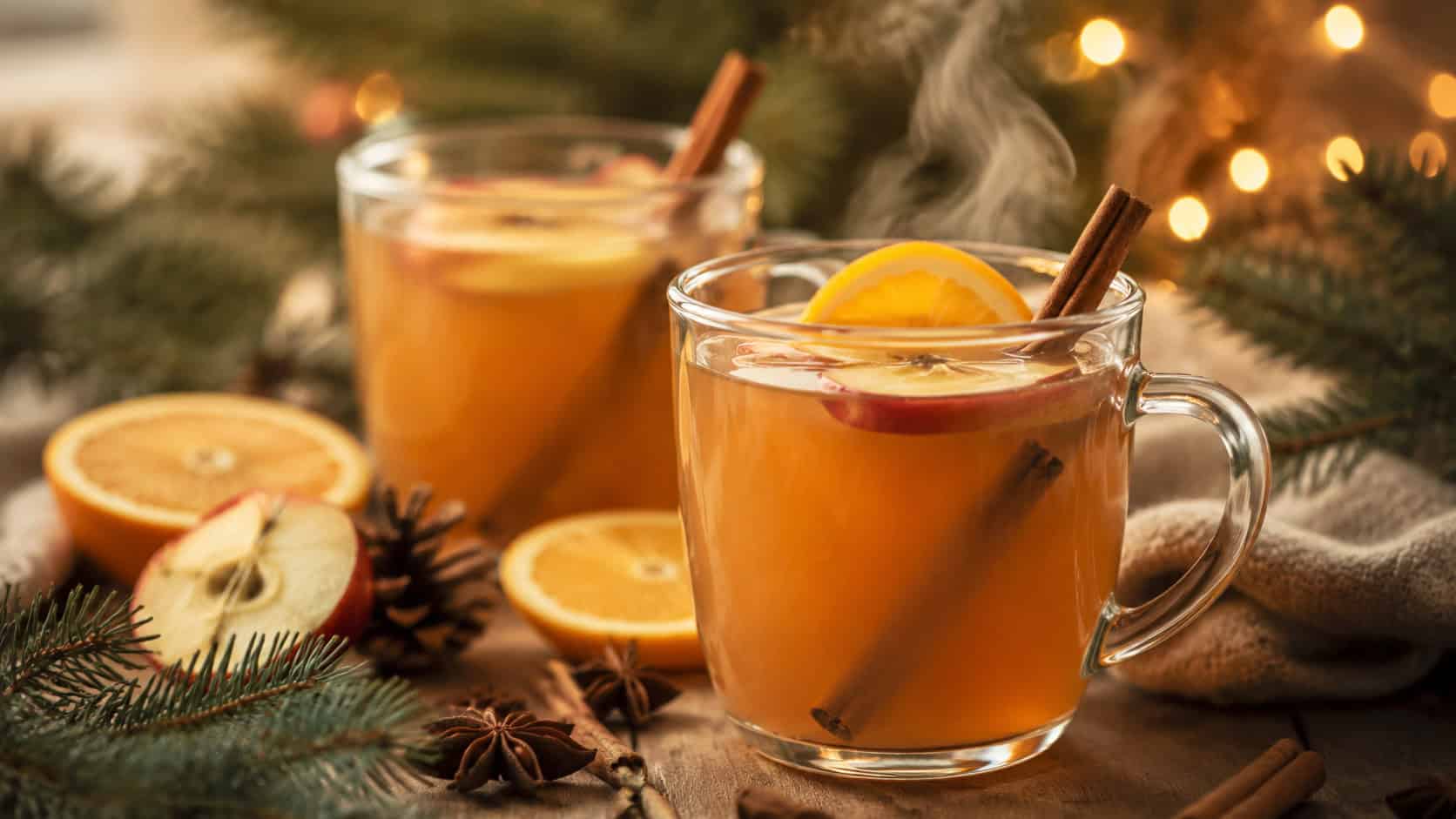 warm spiced apple cider in clear glass mugs garnished with cinnamon sticks and orange slices in a wide horizontal shot, steam rising, set on a cozy table with pine sprigs and twinkling lights blurred behind, natural light, shallow depth of field, landscape orientation
