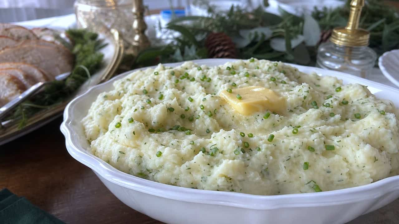 fluffy herbed mashed potatoes in a serving bowl with a pat of melting butter and a scatter of chives in a wide horizontal shot, holiday table with greenery, natural light, shallow depth of field, landscape orientation