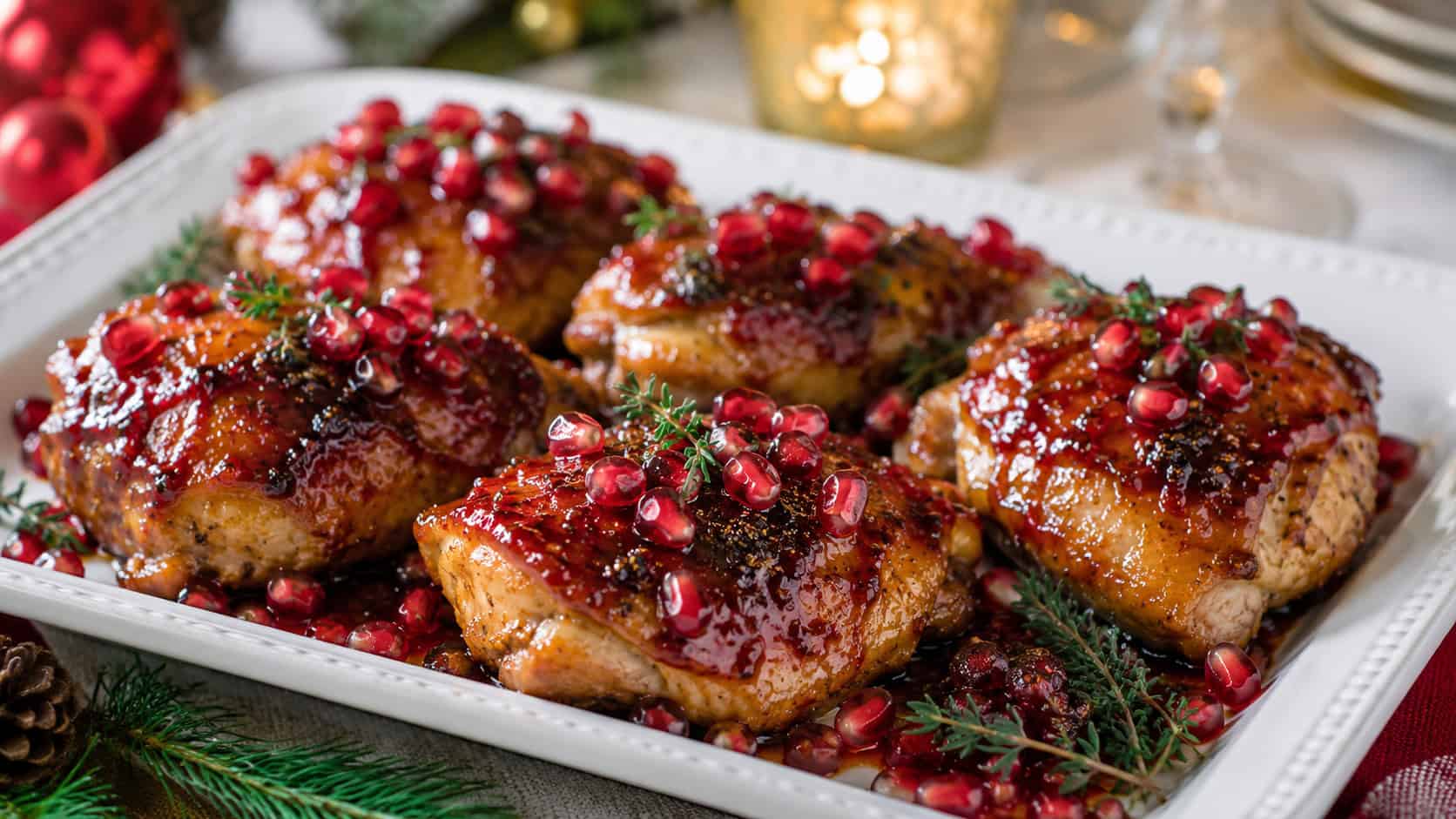 glossy pomegranate-glazed chicken thighs scattered with bright red pomegranate arils and thyme in a wide horizontal shot, on a white platter on a festive holiday table, natural light, shallow depth of field, landscape orientation